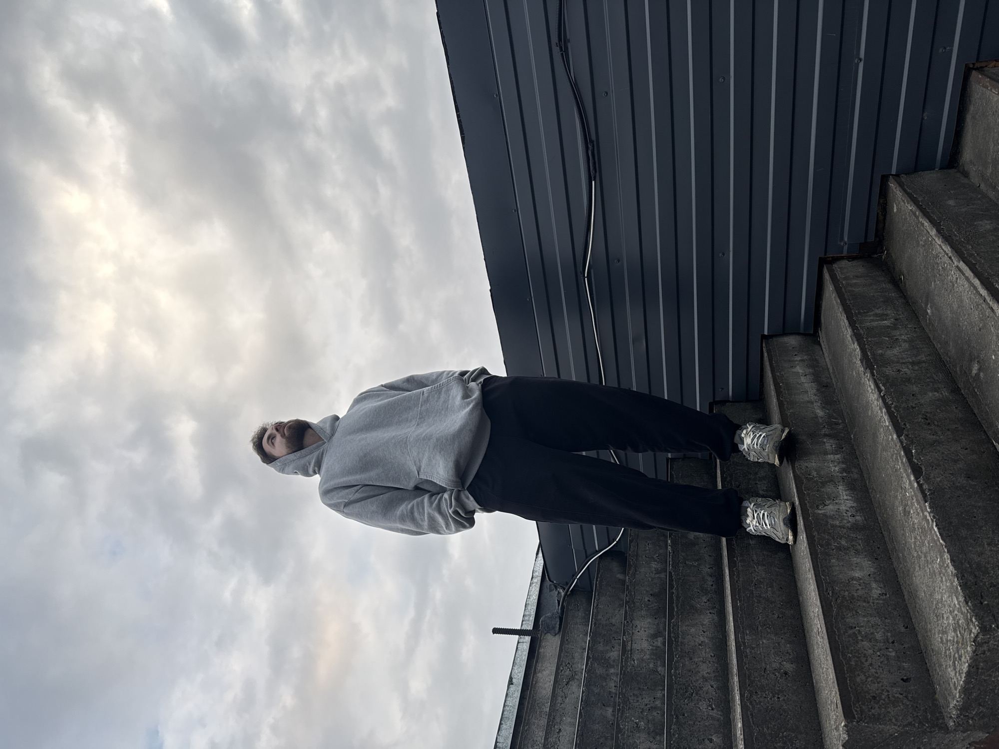 A low-angle shot depicts a man standing on concrete stairs. The man wears a gray hooded sweatshirt and dark-colored sweatpants. He has short brown hair and a beard. The staircase is adjacent to a wall covered in gray corrugated metal siding. The sky, filled with gray clouds, is visible above. The man appears to be looking up towards the sky. The man is wearing grey sneakers. Location: Chaikovsky, Russia.