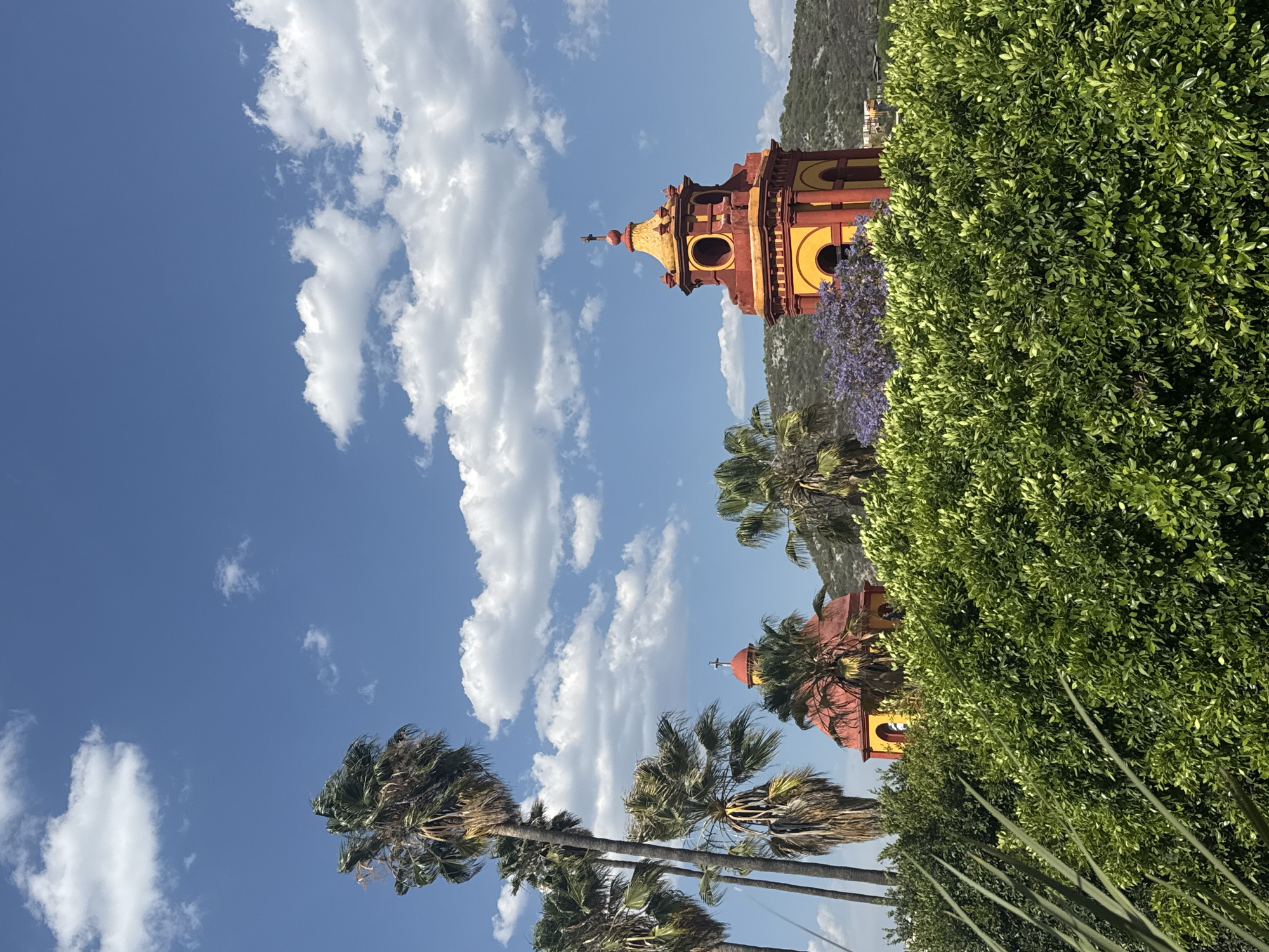 The image shows a view in Bernal, Mexico, with a building featuring red and yellow colors visible among palm trees, greenery, and a blue sky with white clouds. The building appears to be a church tower or bell tower, characterized by its tiered construction, arched openings, and a cross atop the structure. The vegetation includes both dense bushes and tall palm trees. The sky is predominantly blue with numerous white cumulus clouds. The composition suggests a location with a mix of natural and architectural elements, likely taken during a sunny day in Bernal, Mexico.