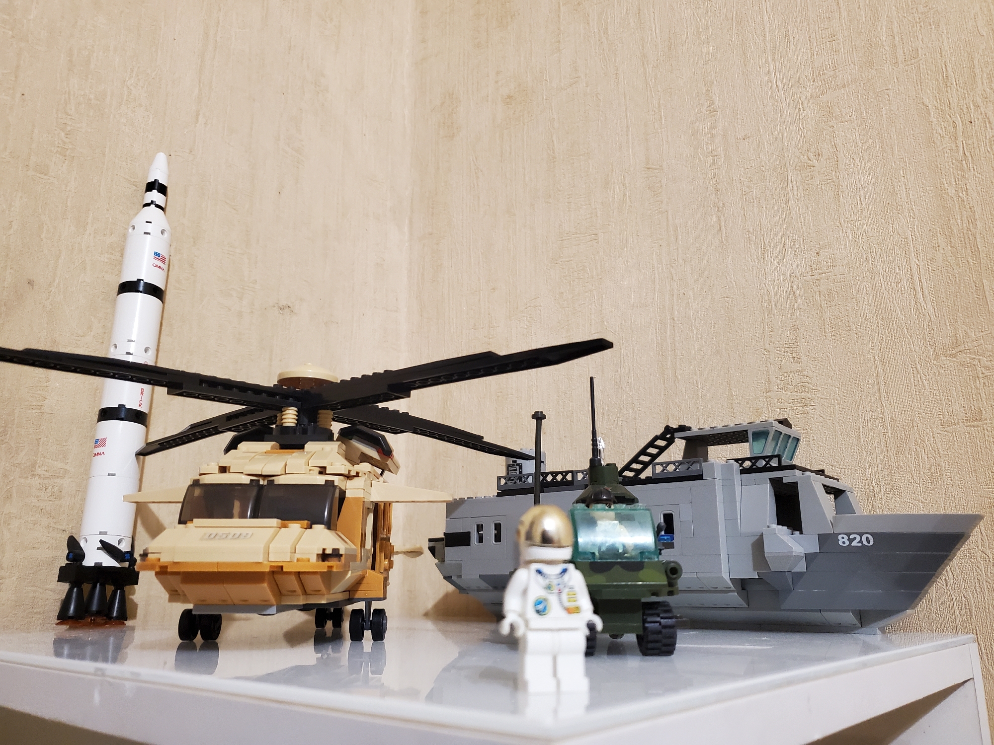 An indoor, eye-level shot captures a collection of LEGO models on a white shelf against a textured, beige wall. The foreground features a white LEGO astronaut with a gold helmet, standing between two helicopters. To the left, a tan-colored helicopter is positioned next to a white rocket with black accents and a US flag decal. To the right, a grey LEGO boat with the number "820" on its hull is visible. A smaller, green camouflage helicopter with black wheels is situated in front of the boat. The lighting is even, and the background wall has a subtle vertical texture.