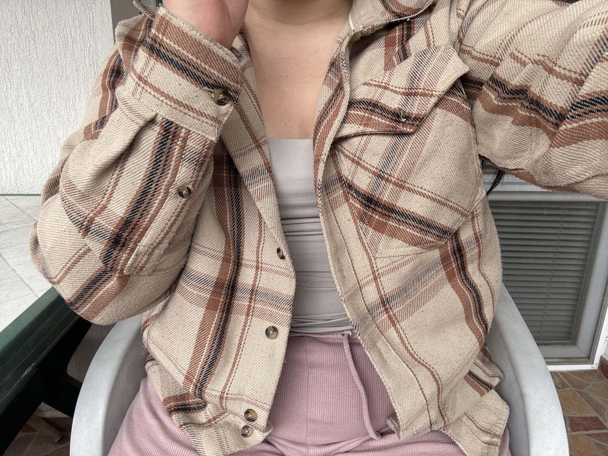 A person is sitting in a white plastic chair, wearing a tan and brown plaid flannel shirt over a gray tank top, and pink ribbed sweatpants with a drawstring. The person is partially visible from the chest up, with one hand near their face. The background includes a green metal frame, a window with blinds, and tiled flooring. The location is likely a balcony or patio. The city is Asenovgrad, Bulgaria.