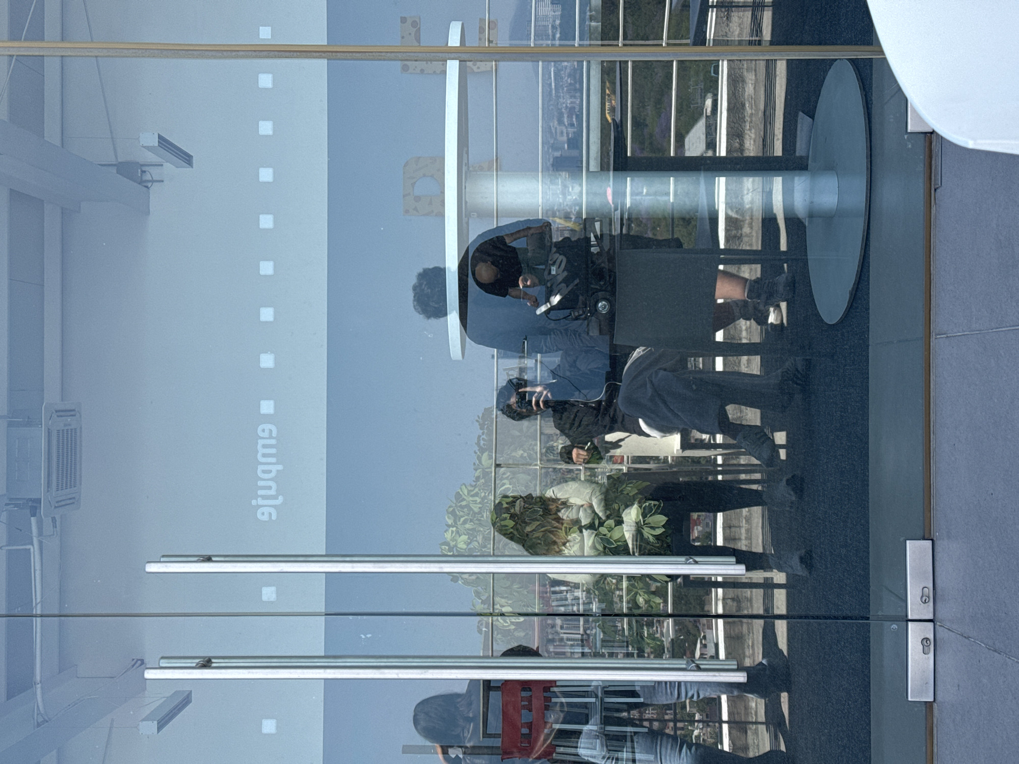 The image depicts a scene observed through a large glass door or window, offering a view of an outdoor terrace against a cityscape background, while simultaneously reflecting elements from the interior space where the photograph was taken. The location is Unknown, Unknown.
**Interior Reflections:**
From the interior, three individuals are reflected. On the far left, a person with long dark hair is seen from behind, standing, wearing a dark top and light pants, with a red object (possibly a chair or crate) partially visible behind them. In the center-left, a seated person, wearing a dark jacket and trousers, is reflected holding a black camera or smartphone to their face, seemingly taking a picture. On the right, a person with short dark hair or bald, wearing a dark short-sleeved shirt, is reflected leaning forward over a piece of black electronic equipment, possibly a camera rig or sound mixing console, on a stand. The word "9tuqm9" (mirrored text) is prominently visible on the glass, along with reflections of ceiling structures, an air conditioning unit, and rectangular light fixtures. Two polished vertical door handles bisect the image vertically in the center. The interior floor is visible as a dark, possibly carpeted, surface with metal door hardware at the base.
**Outdoor Terrace Scene (seen through glass):**
The outdoor terrace features at least two individuals. In the center-left, a person is seated, largely obscured by dense green foliage, appearing to have long, wavy hair and wearing a light-colored top with arms crossed. On the right, a person with dark, curly hair stands behind a white, round table with a central pillar base. Dark chairs are visible around the table, some empty. A metal railing runs horizontally along the edge of the terrace. The outdoor floor appears to be a dark, possibly paved or tiled surface.
**Environment:**
Beyond the terrace, a vast urban landscape stretches into the distance, characterized by numerous buildings and structures under a clear blue sky. Mountains or hills are faintly visible on the distant horizon.