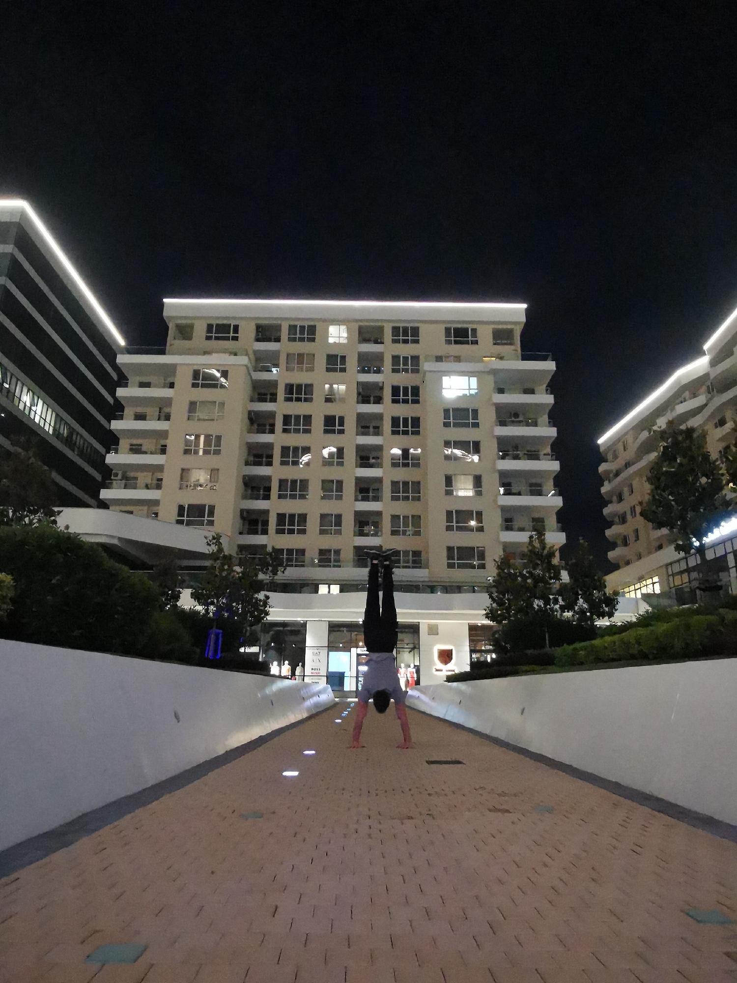 The scene depicts an urban environment at night in Burgas, Bulgaria. A single individual is captured in the center of the frame, performing a handstand on a light-colored paved walkway. The person is dressed in dark trousers and a light-toned (possibly grey) top; their gender is not clearly discernible due to the inverted posture. Their hands are placed on the textured, brick-patterned pavement, and their legs are extended vertically upwards.
The walkway, which recedes into the background, is illuminated by several small, circular, embedded ground lights. It is flanked on both sides by low, white concrete walls, behind which green foliage and small trees are visible.
In the background, three multi-story buildings dominate the cityscape. The central building is a tall, light-beige structure with numerous illuminated windows and balconies across its many floors. Its roofline is accentuated by a bright, continuous white strip light. To the left, a portion of a darker, possibly charcoal-grey building with distinct horizontal white strip lighting is visible. To the right, another light-colored building, similar in architectural style to the central one with balconies and a white strip light along its upper edge, is present. The ground level of these buildings features commercial establishments with illuminated storefronts and visible branding, including partially discernible logos such as "EA7" and "BOSS." The sky above is dark and clear, indicating nighttime, with artificial lighting being the primary source of illumination for the entire scene.