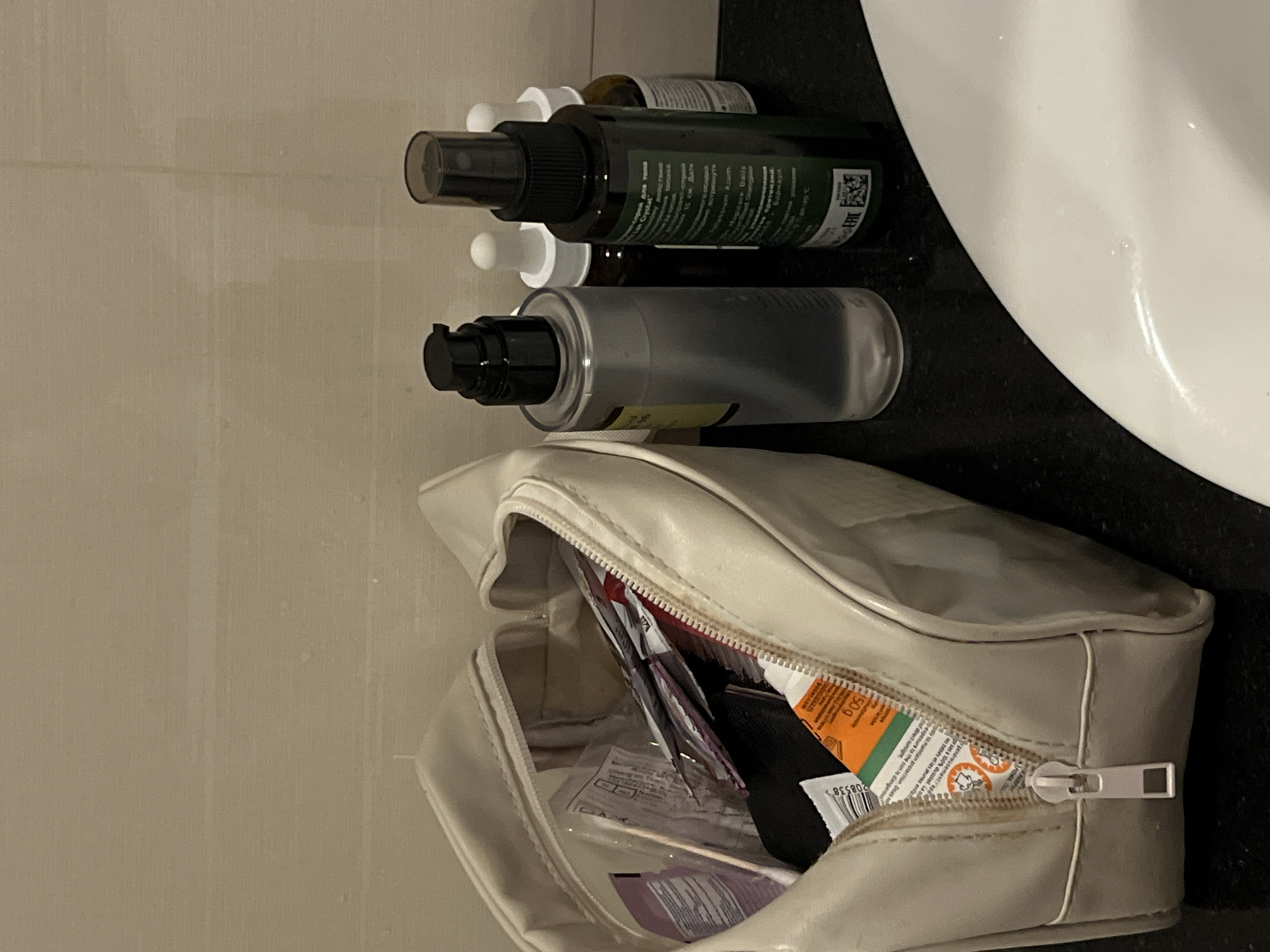 The image displays a collection of personal care items arranged on a black countertop next to a white sink, likely in a bathroom setting within Bac Nha Trang Ward, Vietnam. The scene is shot from a top-down, slightly angled perspective.
In the foreground, a cream-colored toiletry bag with a zipper closure is open, revealing various smaller packets, sachets, and possibly travel-sized items inside. Visible text within the bag includes "150 g" and a barcode-like number "208538>", along with fragmented words like "généreusemanti ap", "les babes et les prunes enfa", and "ysly to maintain protection Deas", suggesting product instructions or descriptions.
Behind the bag, several bottles are neatly lined up. The most prominent is a dark green spray bottle with a black cap, featuring white text on its label, including "ERE" and "To Handwe Daily Crystal", along with some Cyrillic text "15 см. Дать" (15 cm. Give). Next to it is a clear or translucent gray pump bottle with a black dispenser top. Two smaller amber dropper bottles with white caps are partially visible behind these larger bottles.
The countertop is dark, contrasting with the bright white ceramic of the sink basin on the right side of the frame. The background consists of a light-colored, possibly tiled or painted, wall. The overall impression is one of organized personal care products, possibly set up for daily use or unpacking/packing for travel.