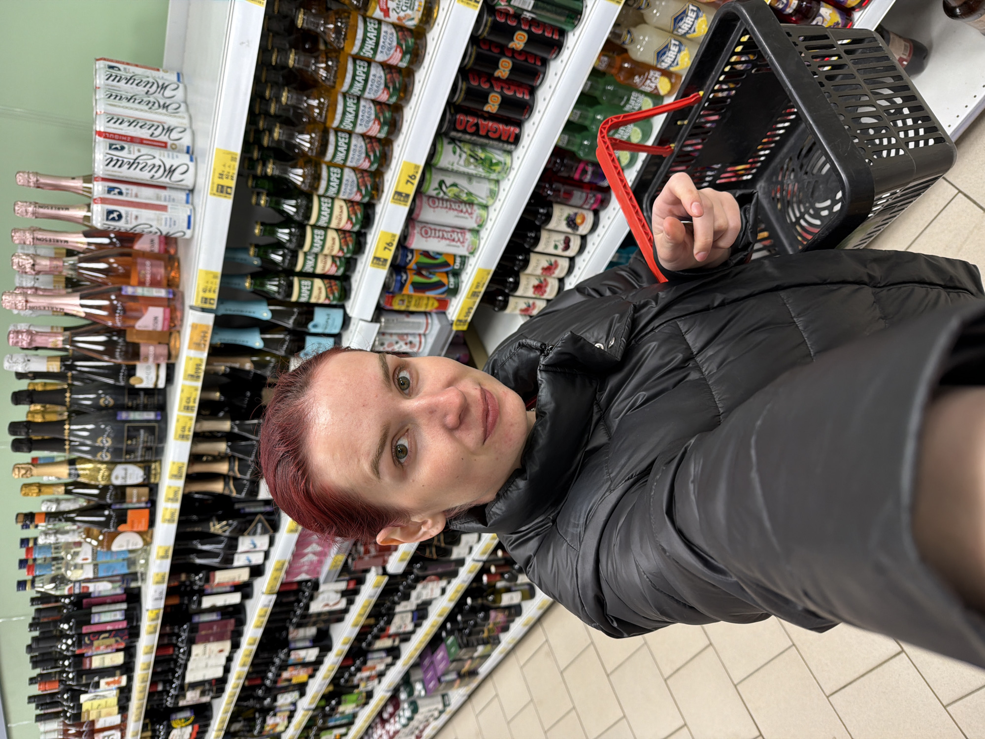 The image captures a selfie of a person, likely in their mid-30s, with red-dyed hair, wearing a black puffer jacket, standing in the liquor aisle of a supermarket. The shelves are stocked with various alcoholic beverages, including beer, wine, and spirits, with labels in Russian and English. The shelves are organized in tiers, displaying a wide selection of brands and bottle sizes. The person is holding a black shopping basket with a red handle, and the background suggests a typical supermarket setting, with tiled flooring and fluorescent lighting. The location is in the city of Chaikovsky, Russia, due to the Russian lettering on the product labels.