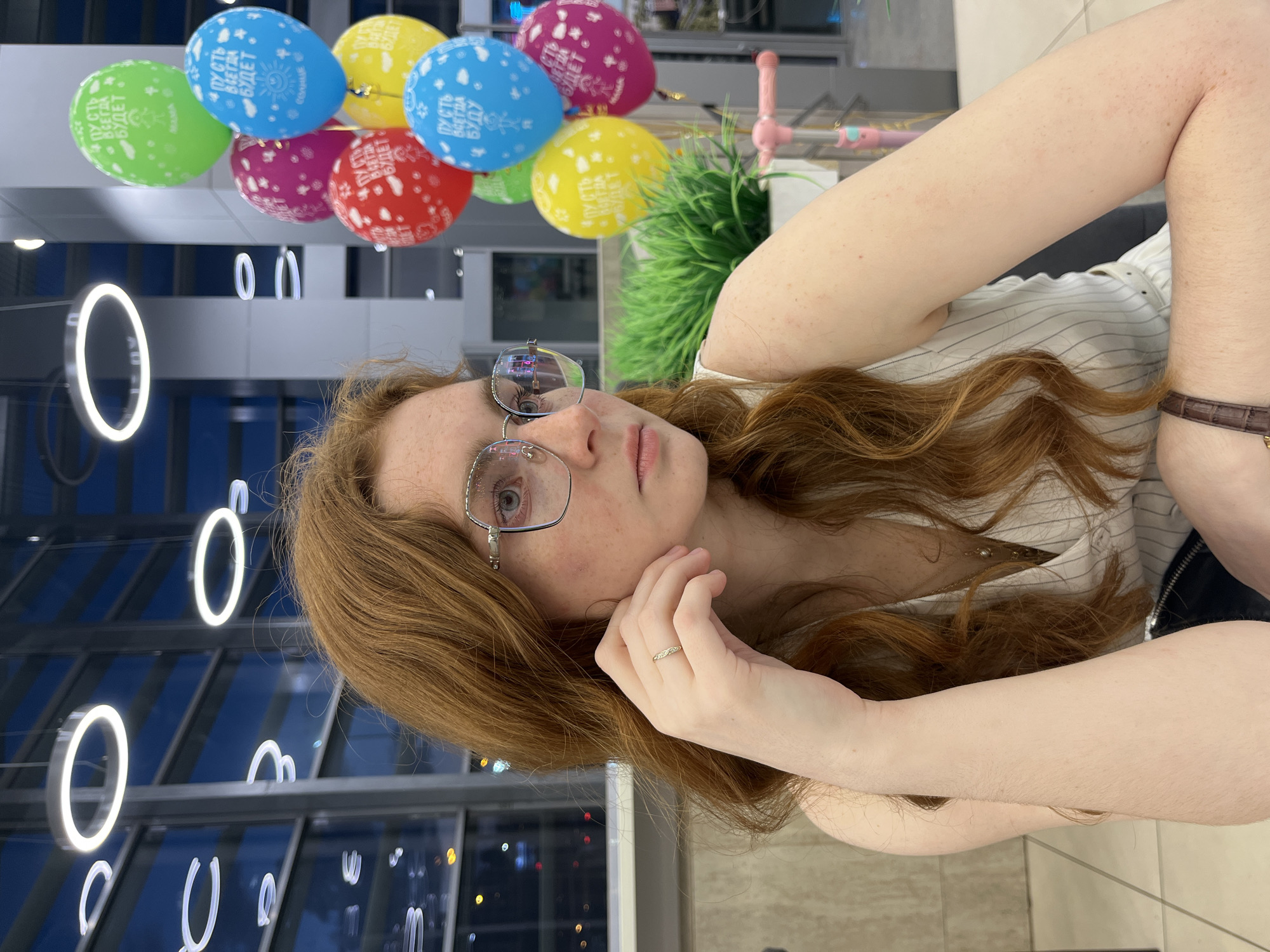 A portrait-style image captures a young woman with long, flowing reddish-brown hair and fair skin, set against a modern indoor background. She is wearing clear-framed glasses and a light-colored top with subtle vertical stripes. Her left hand, adorned with a simple ring on the ring finger, rests gently near her face, while her right arm shows a dark brown watch strap or bracelet. Her gaze is directed towards the camera, with a neutral and slightly pensive expression.
In the upper part of the frame, a cluster of colorful balloons in blue, green, yellow, red, and pink adds a festive touch. Several of these balloons feature Cyrillic text. Clearly visible phrases include "ПУСТЬ ВСЕГДА БУДЕТ" (meaning "LET THERE ALWAYS BE") repeated on various balloons. One blue balloon specifically reads "ПУСТЬ ВСЕГДА БУДЕТ СОЛНЦЕ" (meaning "LET THERE ALWAYS BE SUN"). Another blue balloon has "ПУСТЬ ВСЕГДА БУДЕТ БУДУ", which translates to "LET THERE ALWAYS BE I WILL BE", possibly an incomplete part of a longer phrase or a play on words.
The background reveals a contemporary architectural setting, likely a public space such as a shopping mall or entertainment center, consistent with the provided location context of Kazan, Russia. Distinctive circular white lights illuminate the space. Further back, an artificial green plant and a pink and blue object, possibly a toy or decorative item, are visible. Reflections of distant city lights or indoor lighting are present, suggesting the image was taken during the evening or night. The overall atmosphere conveyed is bright and modern, with a personal moment captured amidst the dynamic environment.