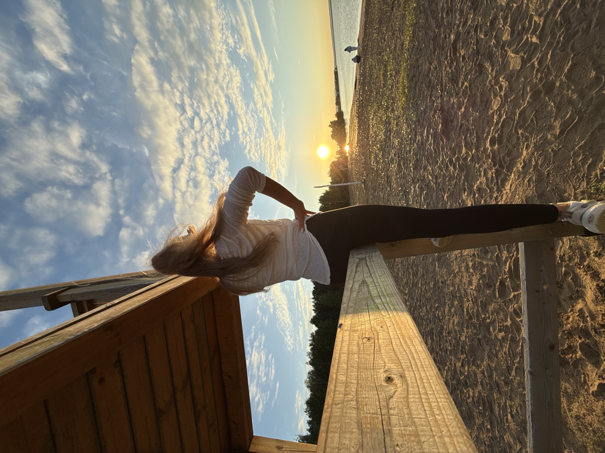 A person with long, light-colored hair, wearing a white long-sleeved top and dark leggings, is seated sideways on a horizontal wooden beam, facing a sunset over a body of water in Chaikovsky, Russia. Their left hand rests on their hip. The wooden beam is part of a larger, multi-component wooden structure, which occupies the left and bottom foreground of the image. A bright, circular sun is positioned low on the horizon over the water, slightly to the left of the image center, casting a golden light across the sandy beach foreground and calm water. Small patches of green vegetation and footprints are visible on the sand. A line of dark trees borders the distant shoreline to the left. The sky features scattered white clouds against a blue background, with some clouds illuminated by the setting sun. Two additional, blurry figures are visible on the sandy beach near the water's edge in the mid-ground to the right. The body of water is identified as the Kama River or the Votkinsk Reservoir, adjacent to the city of Chaikovsky.