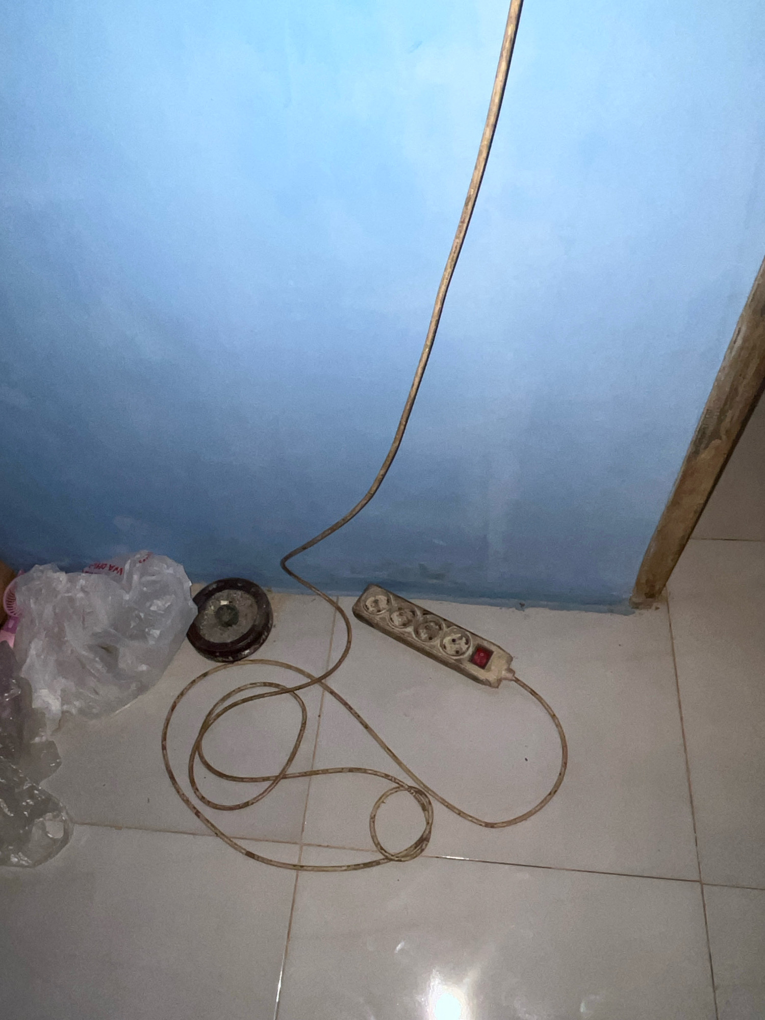 The image depicts a corner of a room with a light blue wall meeting a tiled floor. A light brown electrical cord snakes across the floor, originating from the top right of the frame and leading to a white power strip with four sockets and a red on/off switch. The power strip is located near the bottom right of the image. To the left of the power strip, on the floor, are several objects including a clear plastic bag, a round object with a dark circular center and a metallic rim, and another partially visible object. A thin wooden plank is visible running vertically along the right edge of the frame, adjacent to the wall. The lighting suggests an indoor environment, potentially a residential or commercial space. The specific location in Kebumen, Indonesia is not discernible from the visual information provided in the image.