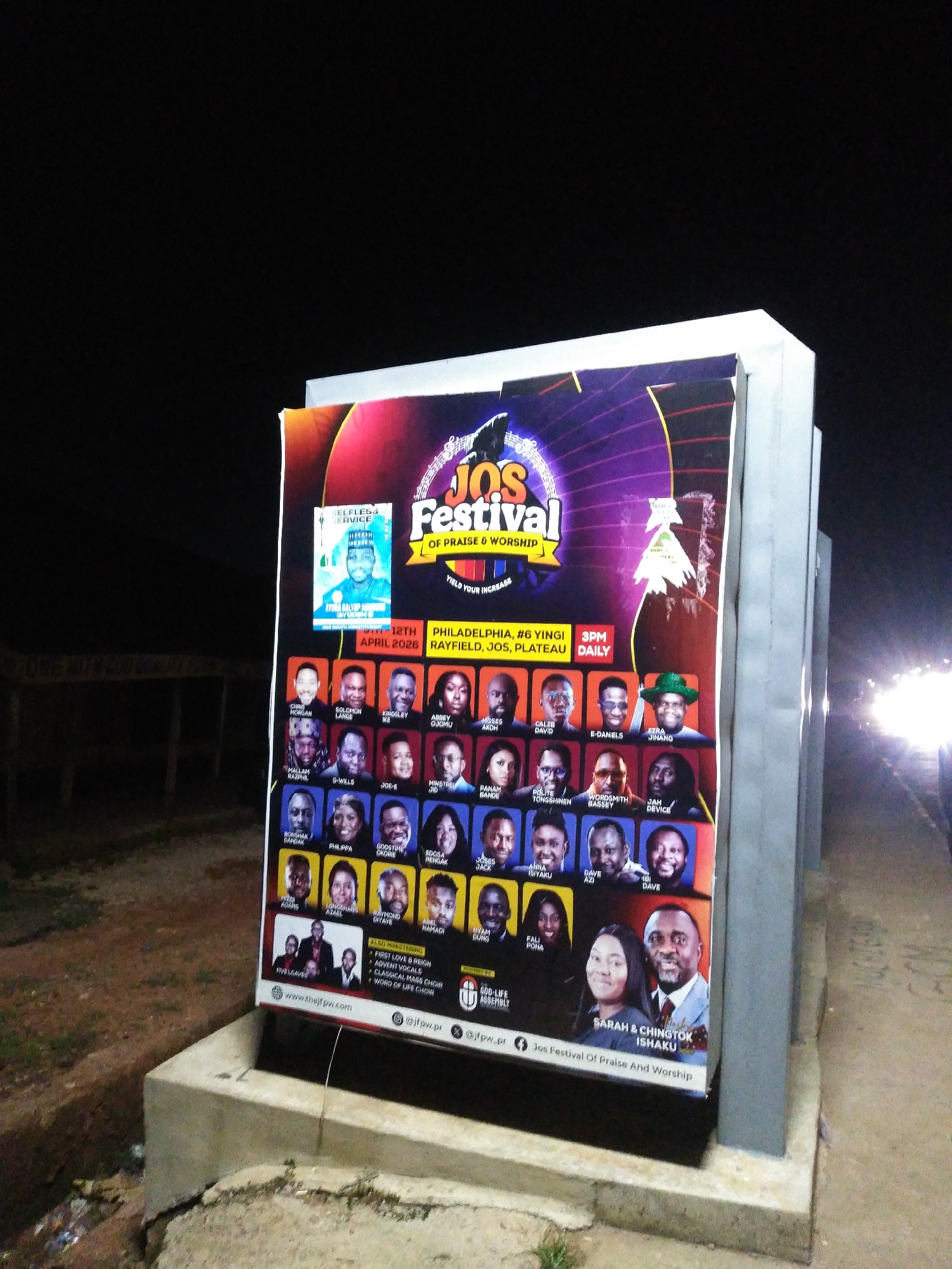 The image shows a large outdoor banner for a "JOS FESTIVAL OF PRAISE & WORSHIP". The banner is illuminated by external lights, and the scene is dark, indicating it is nighttime. The banner features many portraits of individuals, likely performers or speakers, with their names below them. Some of the names visible include "CHRIS MORGAN", "SOLOMON LANGE", "KINGSLEY IKE", "ABBEY OGUJUMU", "MOSES AKOH", "CALEB DAVID", "E-DANIELS", "EZRA JINANG", "BISHOP BAMIDELE", "BANAM BANDE", "POLITE TONGSHINEN", "WORDSMITH BASSEY", "JAH DEVICE", "BONSHAK BANDAK", "PHILIPPA", "GODSTIME OKORIE", "EDOSA HENSARK", "JOEL JID", "SARAH & CHINGTOK ISHAKU", "RYAN DURG", and "FALI POHA".
Text on the banner also indicates the date "APRIL 2026" and locations "PHILADELPHIA, #6 YINGI, RAYFIELD, JOS, PLATEAU". The event is scheduled for "3PM DAILY". Below the portraits, there are smaller sections indicating "ALSO MINISTERING", with listed items like "FIRST LOVE & REIGN", "ADVENT VOCALS", and "CLASSICAL MASS CHOIR". The bottom of the banner displays a website "www.thejfw.com" and social media handles "@jfw.pr" and "@jfw.pr.pr". The banner is mounted on a concrete structure. In the background, there are blurred lights from vehicles, suggesting the banner is located near a road. The surrounding environment appears to be an outdoor space with some vegetation and litter on the ground.
