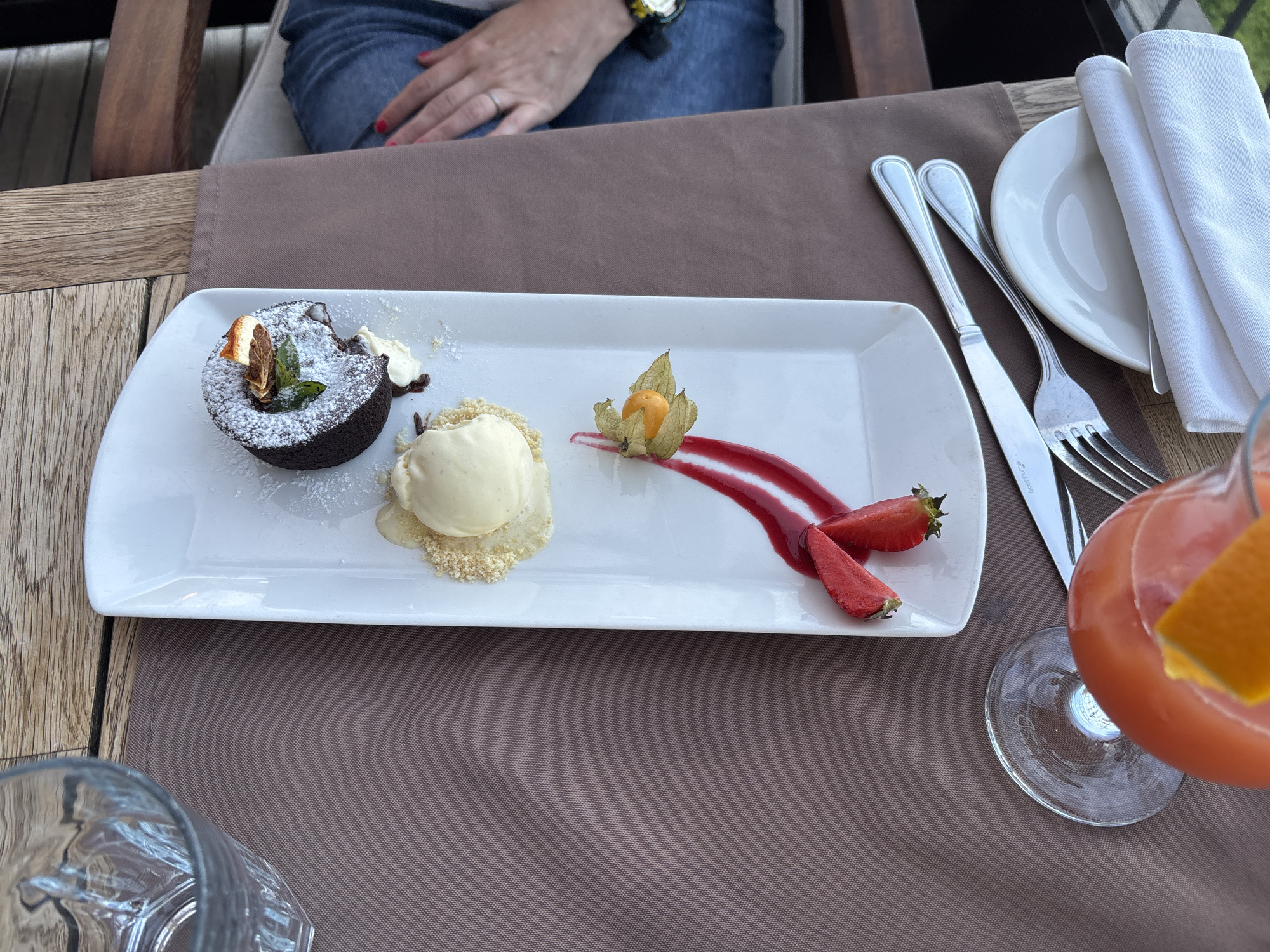 A person, wearing blue denim jeans and a watch on their left wrist, is seated at a table, partially visible in the upper portion of the image. The table is set with a white rectangular plate containing a chocolate lava cake dusted with powdered sugar, a scoop of vanilla ice cream, a halved strawberry, and a physalis. A swirl of red sauce garnishes the plate, and a sprig of mint and a slice of dried orange are on top of the cake. In the background, a white napkin is folded and placed on a white saucer. Silverware, including a knife and fork, is also visible on a brown tablecloth. To the right of the plate, a glass of red-orange liquid with an orange slice garnish is positioned. The scene appears to be set outdoors, as indicated by the wooden furniture and the natural lighting. The context of Topola, Bulgaria, suggests a resort or restaurant setting within that location.