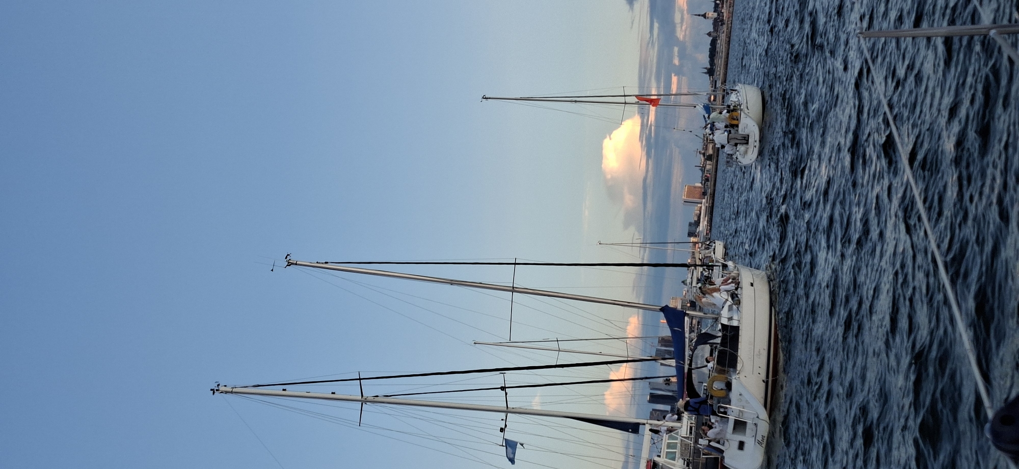 This is a vertical, eye-level shot of several sailboats on the water in front of the city of Tallinn, Estonia. The sky is a light blue with some pinkish-orange clouds, suggesting it is either sunrise or sunset. The water is dark blue with visible ripples, indicating a gentle breeze.
In the foreground, on the left, is a white sailboat with a blue jib sail partially furled. Several people are visible on its deck, appearing to be engaged in activities related to sailing or enjoying the view. Further back and to the right, a second white sailboat is prominent. It has a single red flag flying from its mast, and a group of people are gathered on its stern. The hull of this boat is visible with the name "Amore" on the stern.
Beyond the sailboats, the Tallinn skyline is visible. It features various buildings, including some tall structures and older, possibly historic architecture, suggesting a mix of modern and old city elements. The overall scene depicts a leisurely day on the water with sailboats in front of the city.