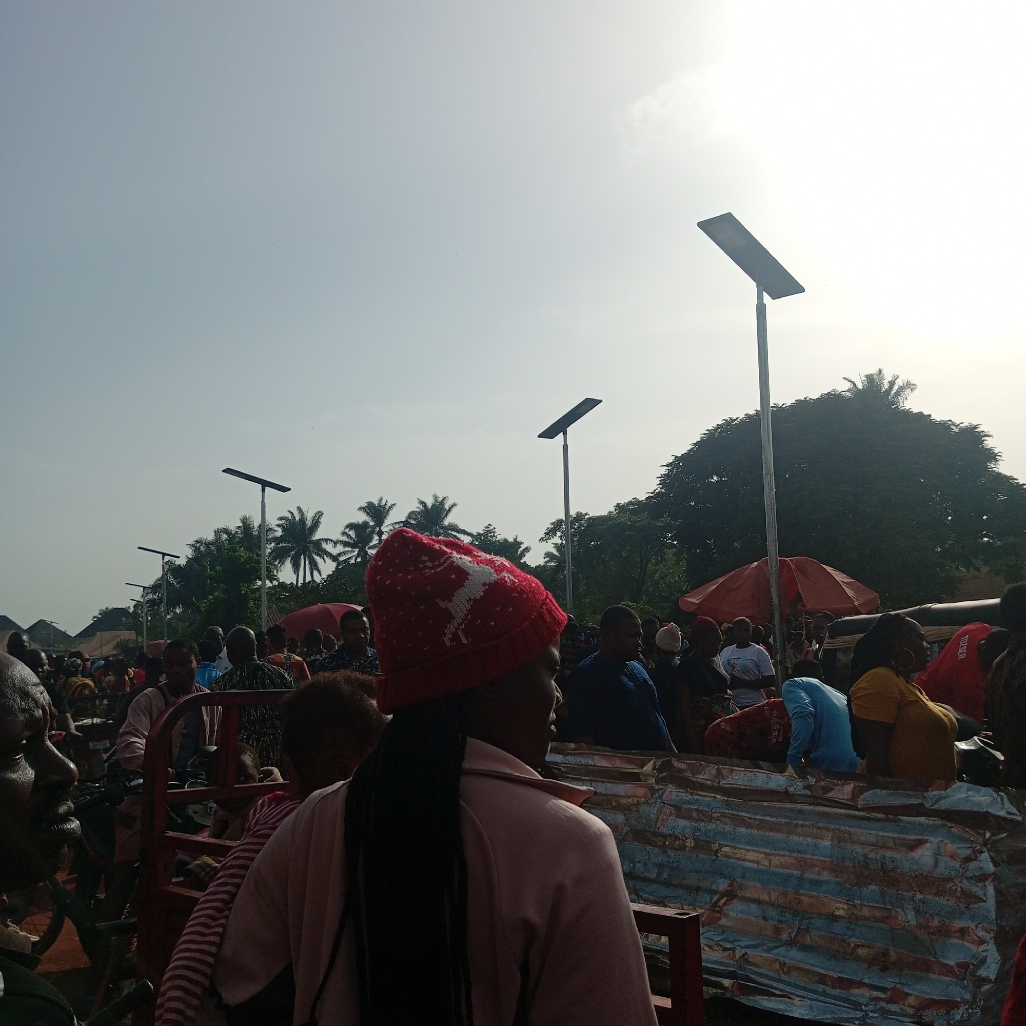 A densely populated outdoor scene in Nsukka, Nigeria, depicts a gathering of numerous individuals amidst market stalls and streetlights. The subjects are primarily African, with a variety of clothing styles and ages represented. Many individuals appear to be engaged in shopping or conversation. In the foreground, a woman wearing a red knit cap stands out. Several solar-powered streetlights are visible throughout the scene. Other trees are visible in the middle of the frame.