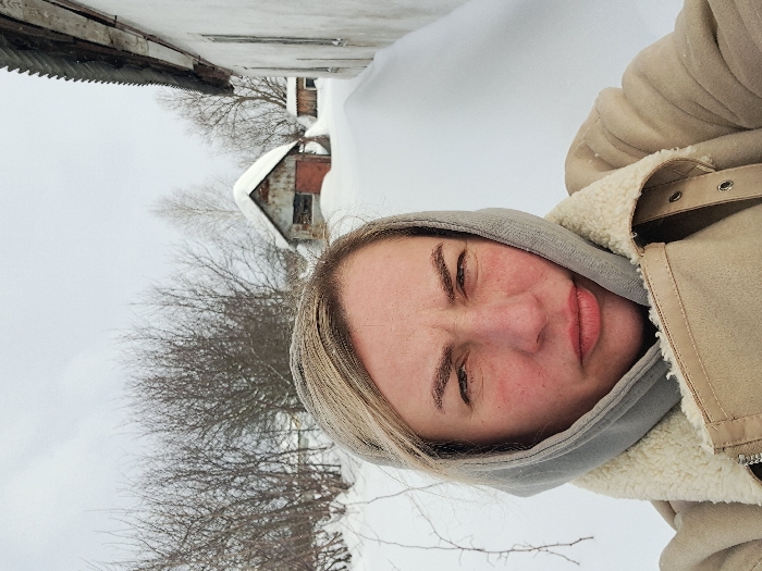 A selfie taken in the city of Полянки, Russia, depicts an adult female in a snow-covered outdoor environment. The subject occupies the foreground, looking directly at the camera with furrowed eyebrows and slightly pursed lips, suggesting a disgruntled or cold expression. Her cheeks exhibit a reddish hue. She has light-colored hair, partially visible from beneath a light grey hood or scarf. She is dressed in a light tan/beige jacket with a visible cream-colored sherpa or fleece lining around the collar. The jacket features a zipper and snap closures. The background is dominated by a winter scene under an overcast sky, with the ground entirely covered in deep white snow. Bare deciduous trees and bushes are visible in the middle ground. To the immediate right of the subject, a section of a white or light-colored building with a dark, corrugated roof is partially visible. Further in the background, a small, weathered shed or outbuilding with a steeply pitched, snow-covered corrugated metal roof and a reddish-brown door is present. A smaller, partially obscured structure stands further behind the primary shed.