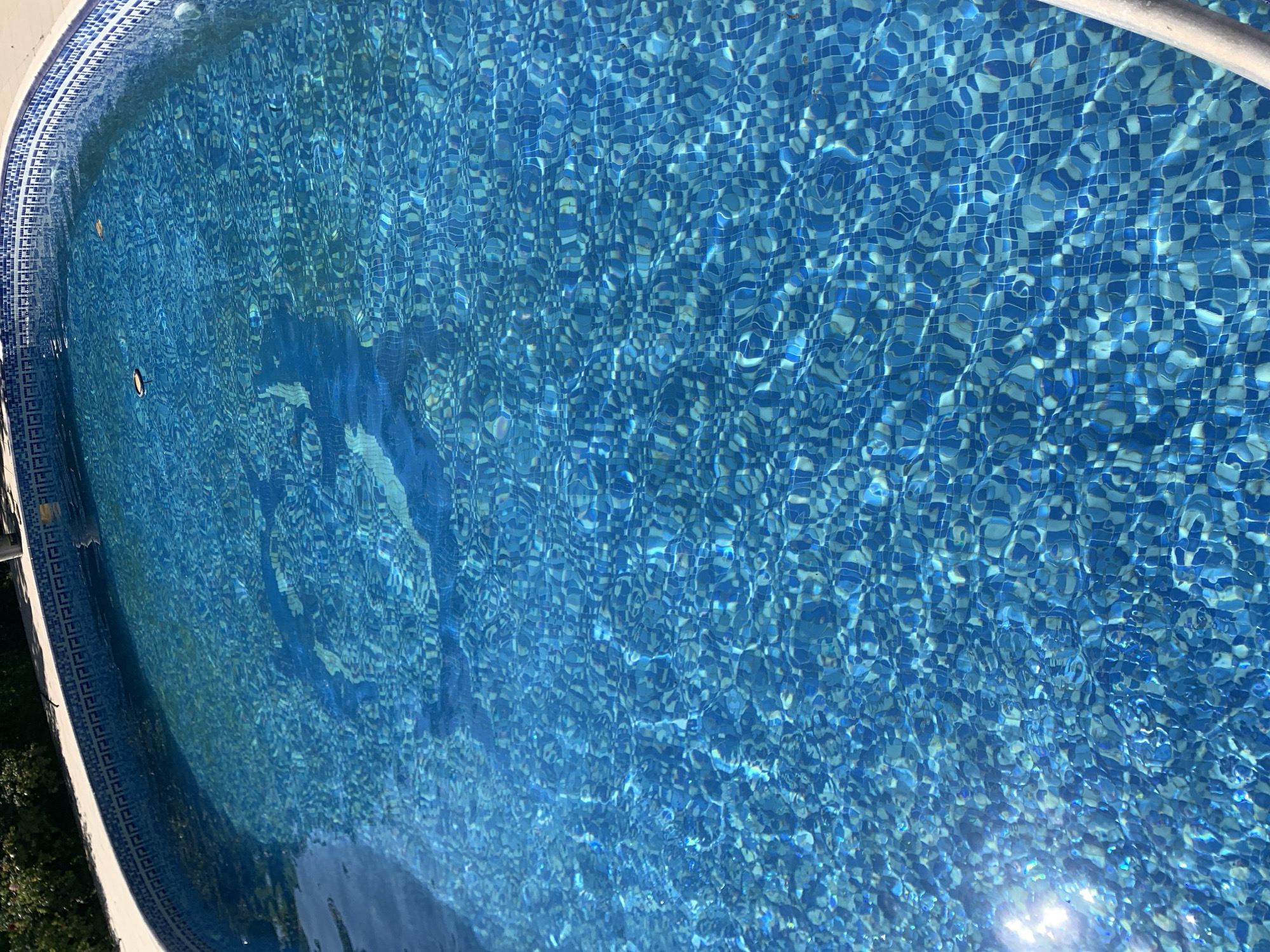 The image shows an above-ground swimming pool with a blue tiled interior. The water is clear and reflects the sunlight, creating ripples and distortions on the surface. The pool is surrounded by a light-colored deck, and a green bush is visible in the upper left corner. The scene is outdoors, suggesting it is a warm day. There are no people or animals visible in the image. The location is stated to be in the Sabunchu Raion of Azerbaijan.
