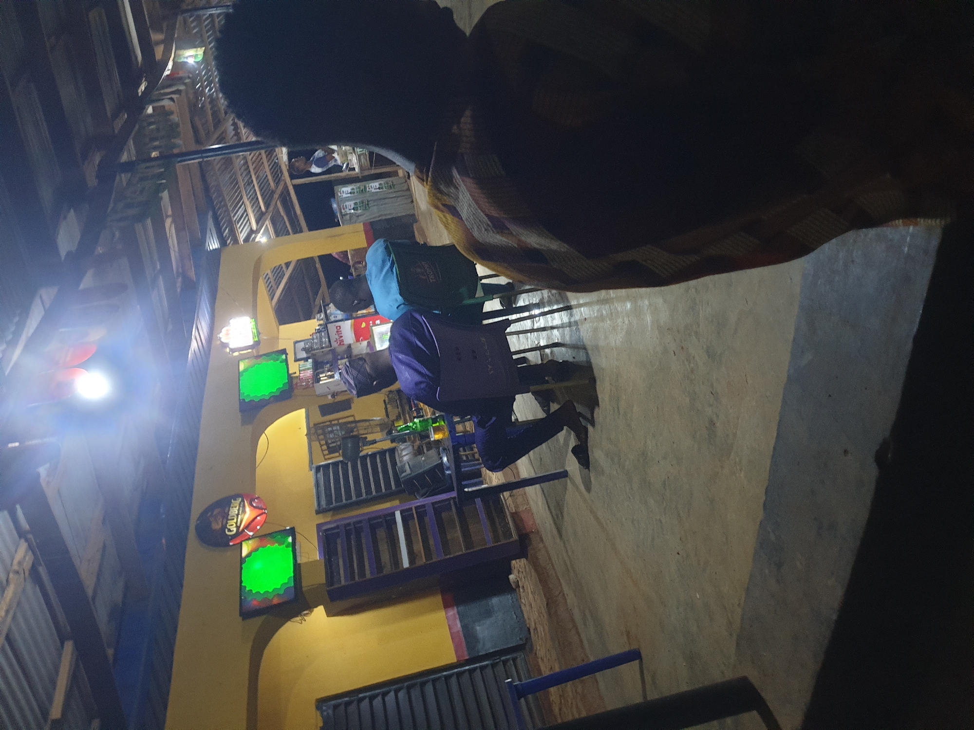 The image depicts a dimly lit indoor/outdoor setting, possibly a bar or open-air establishment in Anilelerin, Nigeria. The floor appears to be concrete and the ceiling is constructed with corrugated metal sheets supported by wooden beams, suggesting a rustic or unfinished construction. In the background, two televisions are mounted on a yellow wall, both displaying bright green screens, indicating they are either off or displaying a blank image. Above these televisions, a circular sign with the "GOLDBERG" logo is illuminated.
To the left, several dark, slatted shutters or doors are visible. In the mid-ground, a table with a bottle of beer and a glass sits between two chairs. A person wearing a purple traditional outfit and a cap is seated in a purple chair at the table, facing away from the camera. Another person, wearing a blue shirt, is seated in a green chair to the right of the first person, also facing away from the camera. A third person, seen from the back and partially obscured by the foreground, wears a patterned garment in shades of orange and brown. In the background, towards the right, another individual in a white and blue shirt is visible, appearing to be standing behind a counter or serving area.
The overall atmosphere is casual and appears to be evening or nighttime due to the lighting conditions and the visible artificial lights. The location suggests a local gathering place within Anilelerin, Nigeria.