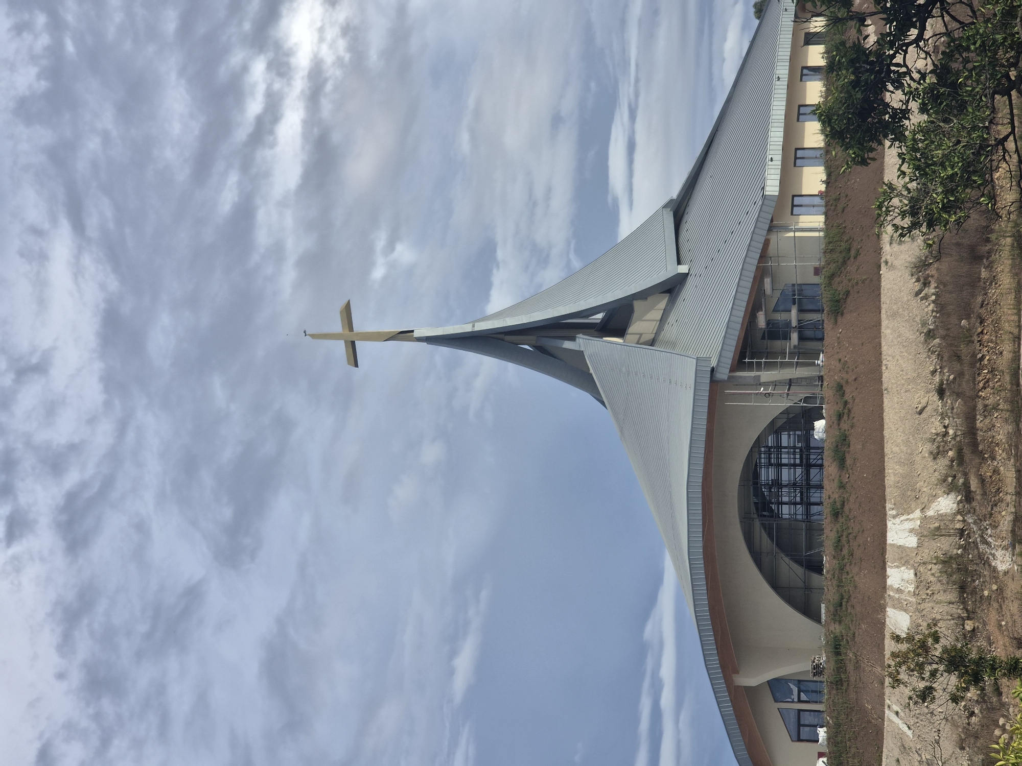The image shows a large, modern building with a striking architectural design, topped with a golden cross. The building appears to be a church or religious institution, with a distinctive, multi-faceted roof structure that culminates in a central spire. The facade features large windows and a light-colored exterior. Scaffolding is visible in some areas, suggesting that the building may be under construction or undergoing renovation. The sky above is filled with dramatic, cloudy formations. The building is situated on a slope, with a natural, somewhat uncultivated landscape in the foreground. This building is the Igreja de Nossa Senhora da Fátima in Póvoa de Varzim, Portugal.