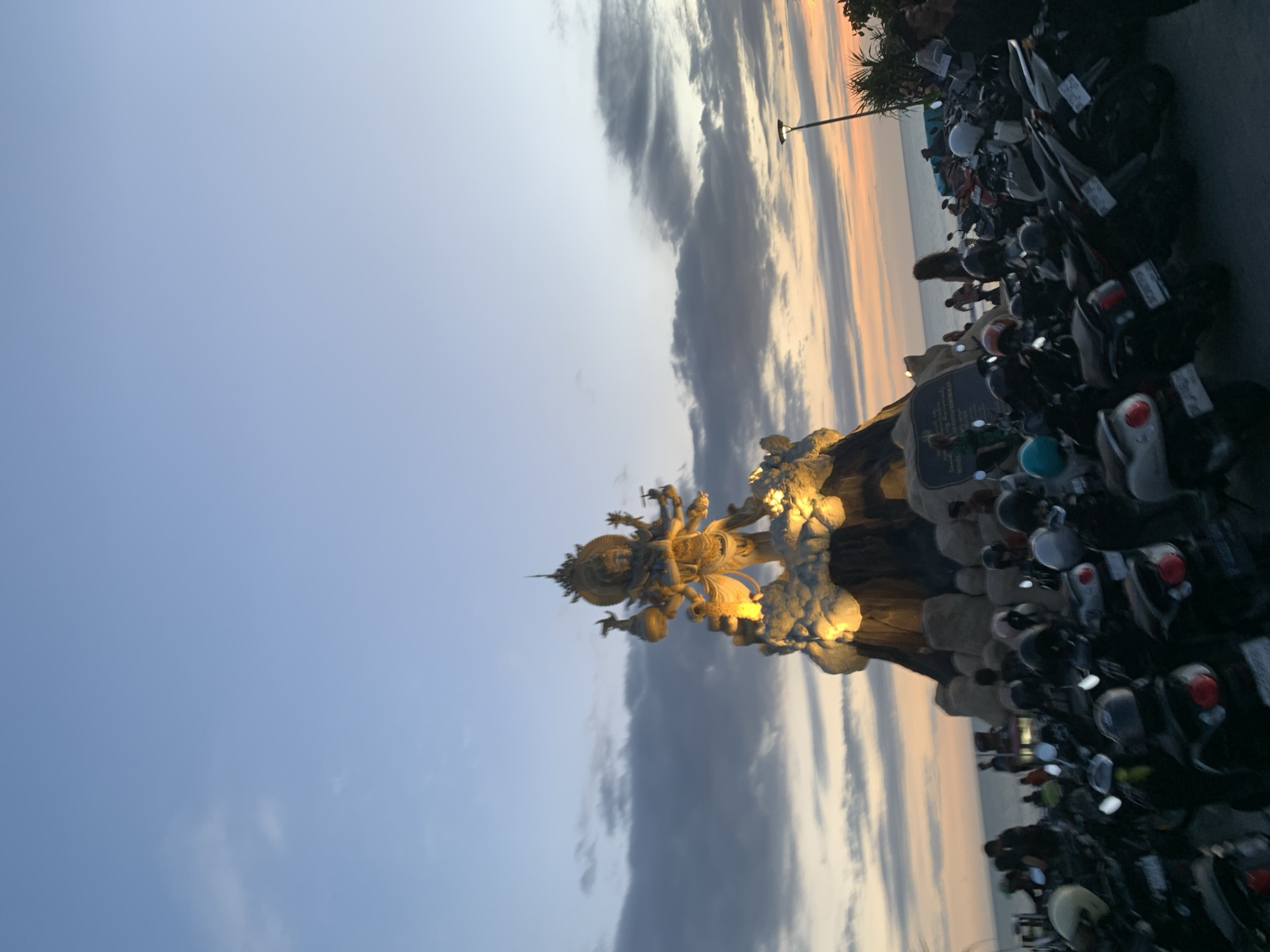Captured at sunset in North Kuta, Indonesia, the image showcases a statue brightly lit, potentially depicting a deity, positioned to the right of the frame. The backdrop features a sky transitioning from blue to orange with bands of grey clouds running vertically. Parked motorcycles fill the lower right quadrant, with a street light visible in the distance. The scene appears to capture a blend of cultural art and the surrounding urban environment during the evening.