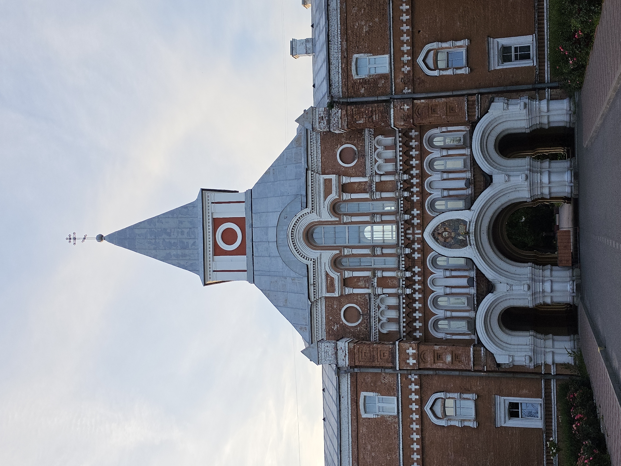The image shows the main entrance of the St. Sergius Orthodox Church in Strelna, a town within the federal city of St. Petersburg, Russia. The church is constructed of red brick and features a prominent bell tower with a spire topped by a cross. The entrance is an arched gateway supported by white stone columns and decorated with ornate carvings. Above the archway, there is a mosaic depicting a religious figure. The surrounding area is landscaped with rose bushes and a paved walkway leading to the entrance. The sky is overcast with some clouds. There are no people visible in the image.