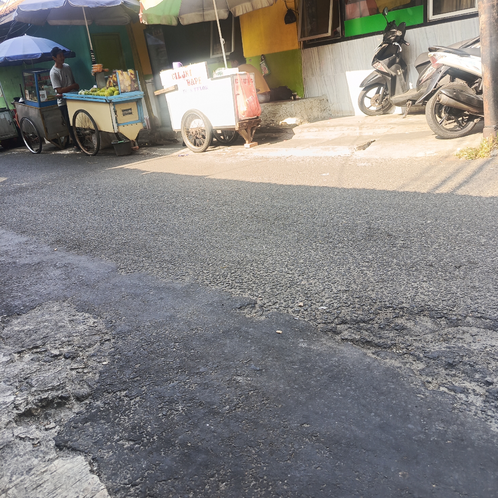 The image depicts a street scene in South Jakarta, Indonesia, with two street vendors and parked motorcycles. In the foreground, a weathered asphalt road shows signs of wear and repair. To the left, a man stands behind a food cart with a blue and yellow canopy, seemingly engaged in selling produce. The cart is equipped with a glass display case and wheels. Further to the right, another vendor is visible near a white cart, also with a canopy, and what appears to be a red plastic bag hanging from it. Two motorcycles, one black and one white, are parked on the sidewalk or paved area to the right of the vendors, against a building with green and yellow walls. The overall atmosphere suggests a typical urban commercial area during daylight hours, with sunlight casting shadows across the road.
