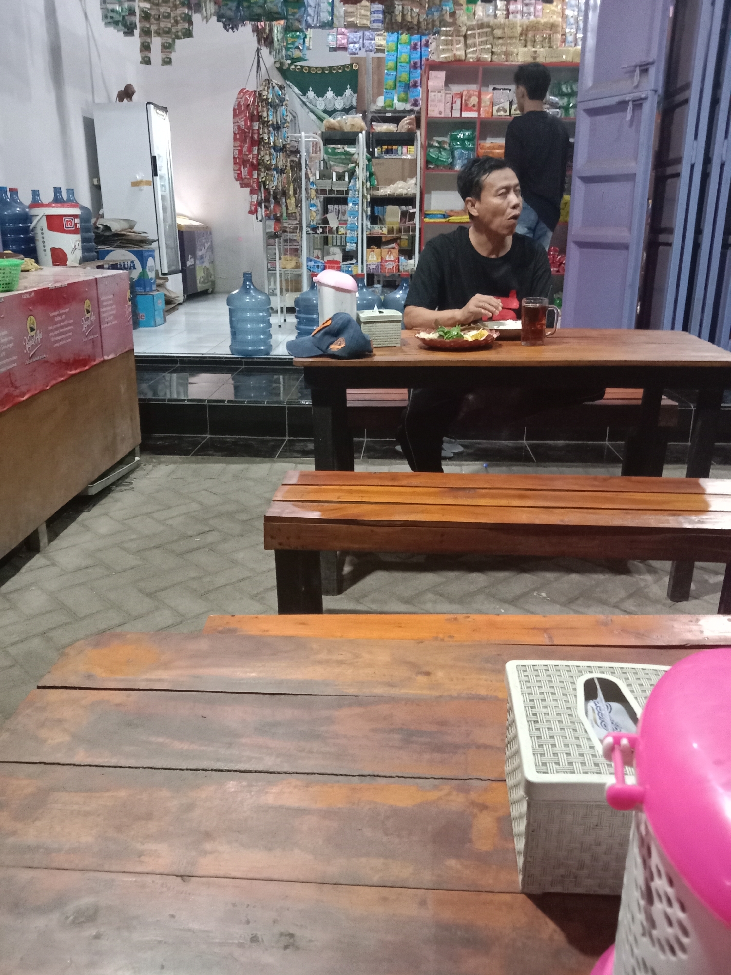 This image captures an indoor scene within a small shop or warung located in Cangkring, Indonesia, appearing to be a local establishment with both retail and dining capabilities.
Two adult males are present. In the midground, an adult male, approximately 30-40 years old with short dark hair, is seated at a rustic wooden table. He is wearing a black t-shirt and has a plate of food (comprising rice, green vegetables, and a dark ingredient) and a clear glass mug containing a dark beverage positioned in front of him. His mouth is slightly open, and his gaze is directed towards the right side of the frame. On the same table, a light-colored rectangular woven tissue box is visible. Behind him, another long wooden bench is present. In the background, a second adult male, viewed from behind, stands near a set of red and white shelves stocked with various packaged goods. He wears a dark t-shirt and light-colored trousers, seemingly engaging with the merchandise.
The shop interior features a retail section on the left, containing multiple large blue plastic water dispenser bottles, a white standing refrigerator/freezer with a small lion figurine on its top, a smaller display cooler, a red and white plastic bucket, and several cardboard boxes. A red counter with visible "Kopi API" branding partially extends into the foreground. The right side of the shop displays extensive red and white shelving units filled with numerous packaged items, including snacks and beverages. Various smaller packaged products are suspended from the ceiling by strings, alongside a decorative green textile with a scalloped edge. A purple door or partition is visible on the far right, beyond which dark metal grating or a shutter is present.
The floor in the seating area is constructed from gray interlocking pavers, transitioning to a darker, possibly tiled, surface in the shop area, delineated by a black step. The walls are primarily white. The entire scene is illuminated by artificial lighting, indicating an indoor or evening setting. The seated male appears to be pausing during his meal, potentially reacting to or conversing with someone or something off-camera, while the standing male is occupied with the shop's inventory.