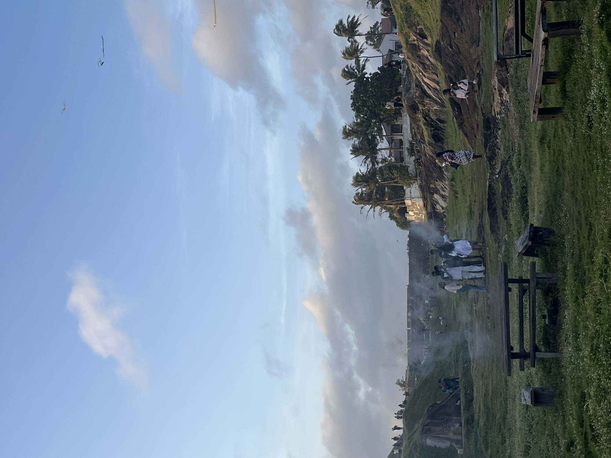 The image depicts a scene at Galle Fort, Sri Lanka, during what appears to be a late afternoon or early evening. The foreground features a grassy area with two wooden picnic tables and benches, some of which are partially obscured by the surrounding foliage. Smoke rises from a fire in the middle ground, where a group of people are gathered. The middle ground also includes a grassy slope leading up to a fortified wall, with more people visible along the top. In the background, palm trees and buildings are visible on a hill. The sky is a mixture of blue with scattered clouds. Two kites are flying in the upper portion of the sky.