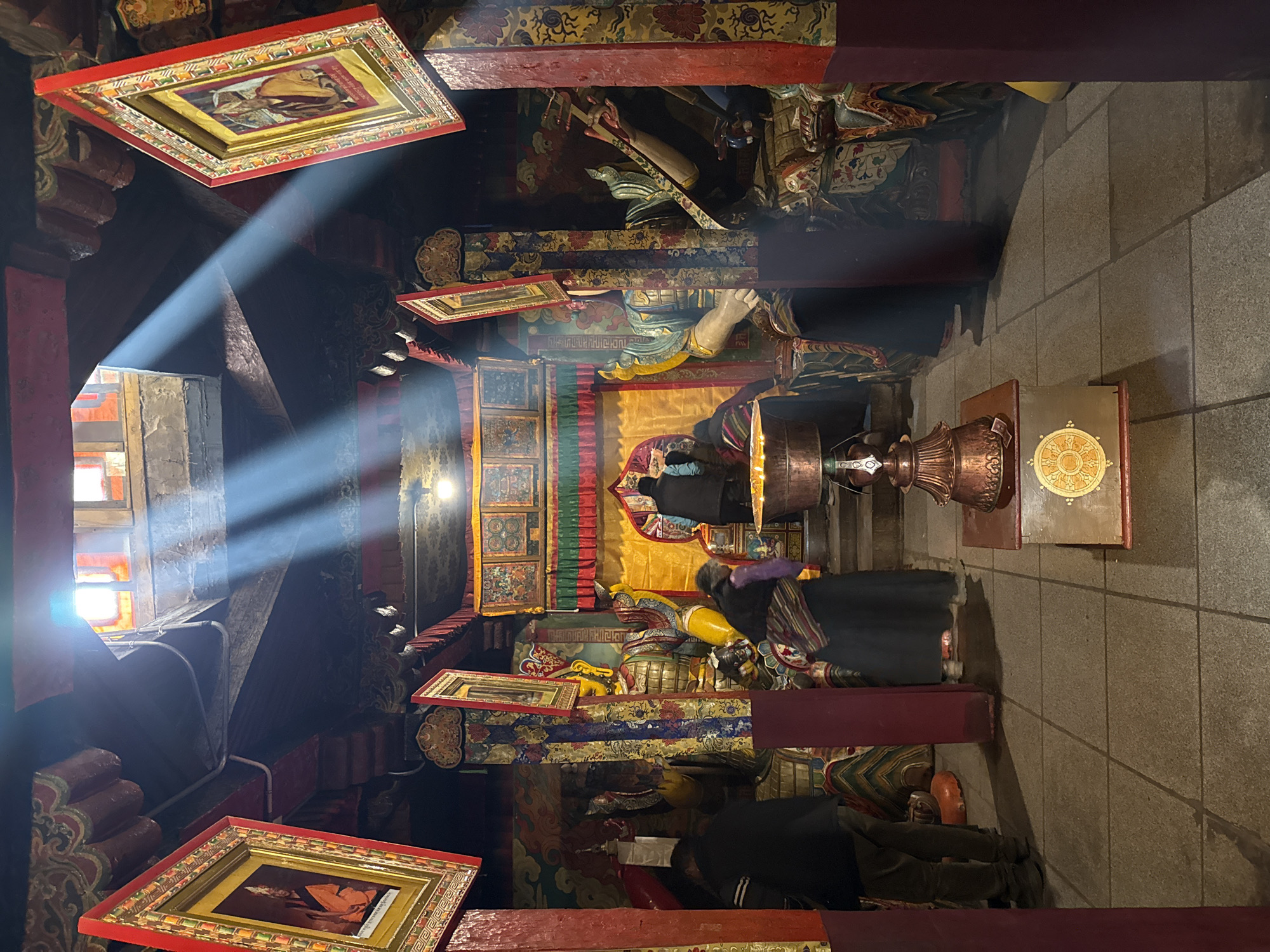 The image captures the interior of what appears to be a Buddhist temple or monastery, characterized by its ornate, traditional design. Dramatic rays of sunlight stream through a window on the left, illuminating dust particles in the air and creating distinct beams across the space. The room features robust wooden beams and pillars, painted in deep reds, gold, and other vibrant colors, adorned with intricate floral and geometric patterns.
Several framed artworks, possibly thangkas or portraits of revered figures, hang prominently from the ceiling and walls, also encased in decorative red and gold frames. In the background, large, colorful statues or figures, resembling guardian deities, are visible, one of which appears to be holding a sword.
In the mid-ground, a person in dark clothing with colorful patterned trim is kneeling or bowing on the stone-tiled floor, facing toward what seems to be an altar or shrine area. Another individual is partially visible kneeling on the far right. A third person is seen further back, also in a posture of reverence. A large, ornate copper-colored vessel or urn rests on a stand near the center, next to another stand holding an object decorated with a golden mandala-like symbol.
The visible text on the framed artworks is too small and indistinct to read clearly, but it appears to be in a script characteristic of Tibetan or similar languages. The scene conveys a strong sense of devotion and spiritual practice, likely taking place in the morning or late afternoon given the angle and intensity of the sunbeams. The architecture and decorative elements suggest a cultural context rooted in Himalayan or Tibetan Buddhist traditions.