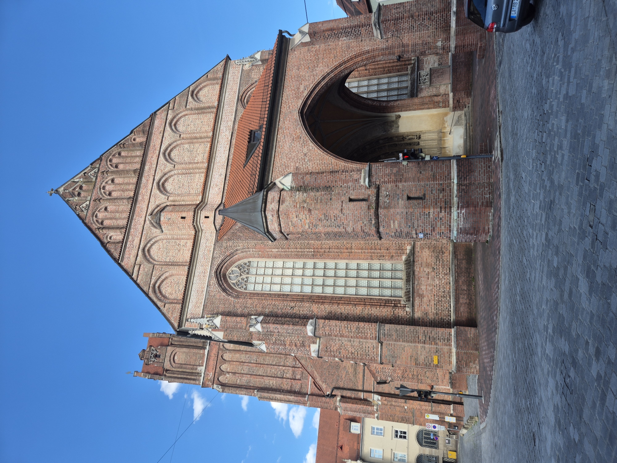 The image shows a large, red brick Gothic church, likely St. Martin's Church, in Landshut, Germany. The church has a tall, narrow tower on the left and a large, arched entrance on the right. The facade is adorned with arched windows and intricate brickwork. The street in front of the church is paved with cobblestones. A grey car is parked on the right side of the street, and a traffic light is visible near the entrance. The sky is blue with a few white clouds.