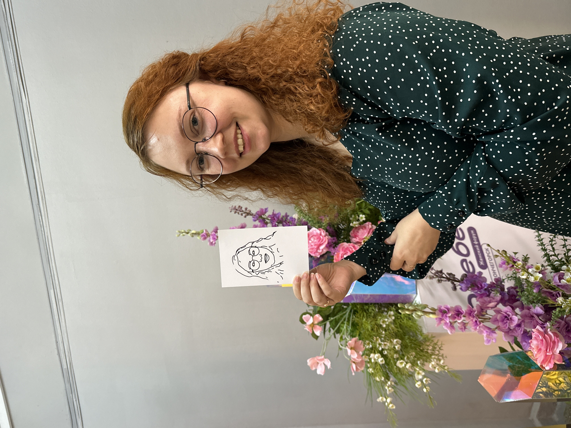 A woman with curly red hair and glasses is smiling and holding up a small drawing of a face. She is wearing a dark green, polka-dotted shirt. Behind her and to the side are pink and purple flowers, greenery, a gift bag, and an iridescent box. The gift bag has text in Russian, specifying "Женский форум" and "МОНЧЕГОРСК," indicating a women's forum in Monchegorsk, Russia. The background is a light grey wall.