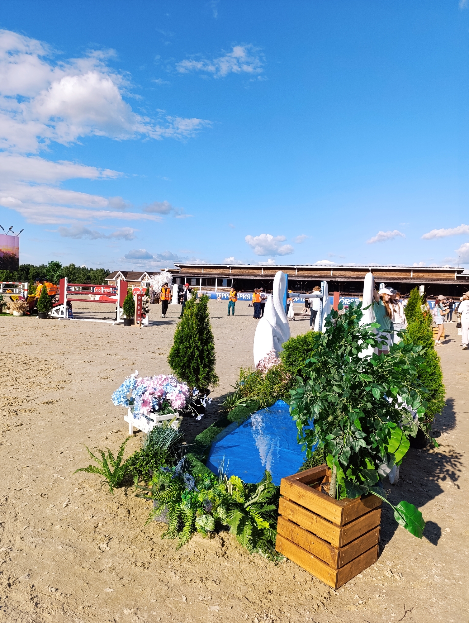 The image depicts an outdoor equestrian event, likely a show jumping competition, in Ivanovskoe, Russia. The foreground is dominated by a decorative setup featuring a small blue pool, artificial plants, and a wooden planter with a large green tree. Behind this, a sandy arena is visible, with several jumps and barriers for the competition.
In the background, a large building with a sloped roof and wooden siding serves as a spectator or event facility. Several people are present, some wearing bright orange vests, possibly staff or participants. The sky is a vibrant blue with scattered white clouds, suggesting a clear and sunny day, likely in the late afternoon due to the shadows. The overall mood seems to be one of active event engagement and pleasant weather.
Visible text includes "AI01" and "HORSE" on various elements of the jumps, along with Cyrillic text on a banner in the background, which appears to be "ТЕРИТОРИЯ СПОРТА" (TERRITORY OF SPORT).