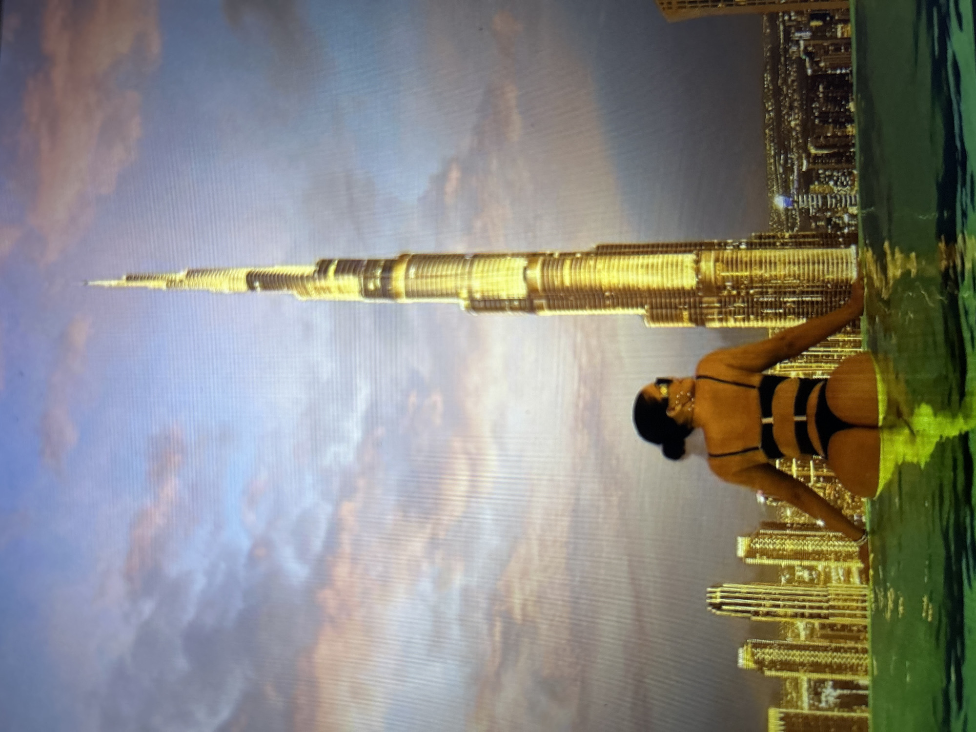 A woman in a black bikini stands in a pool with a cityscape in the background. She is facing away from the camera, with her arms resting on the edge of the pool. The cityscape features several tall buildings, including a prominent skyscraper that appears to be the Burj Khalifa. The sky is cloudy and has a warm, sunset-like glow. The water in the pool is a vibrant green color. The image does not depict the city of Burgas, Bulgaria.