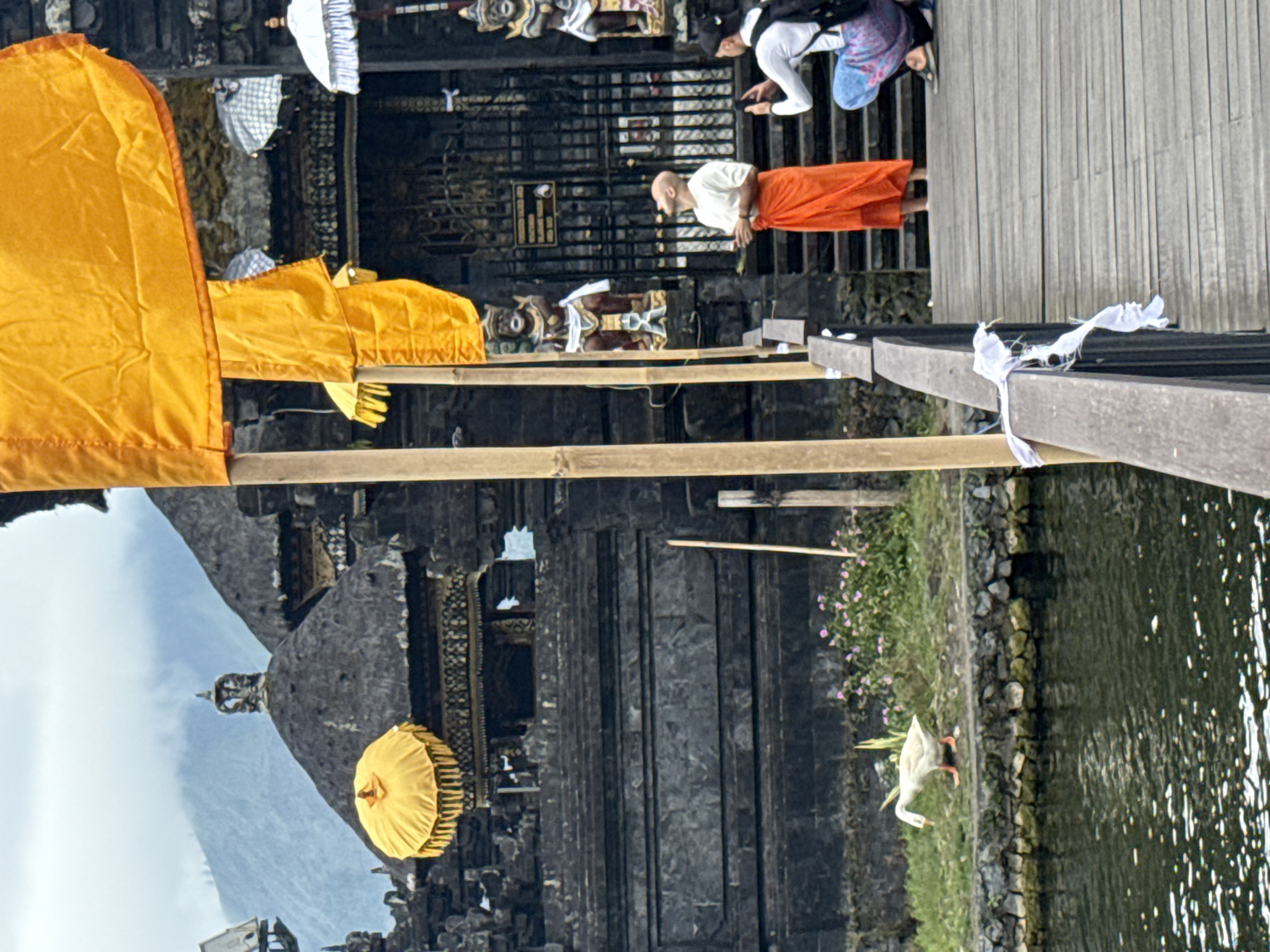 This image captures a scene at a temple complex in Kintamani, Indonesia, with a prominent body of water in the foreground. A wooden walkway extends into the frame from the right, leading towards a stone temple structure with dark, textured walls and tiered roofs. Two orange and yellow decorative umbrellas are visible, one on a rooftop and another hanging from a pole. Colorful flags, primarily orange, flutter from bamboo poles.
In the background, a misty mountain range looms under an overcast sky. On the left side of the frame, a white goose with orange feet walks on a grassy bank bordering the water.
On the wooden walkway, two individuals are present. A person dressed in a white shirt and an orange sarong stands looking upwards, holding what appears to be a small bunch of leaves. To their right, another person kneels, taking a photograph with a smartphone. A backpack is visible on this person's back. The walkway is adorned with white prayer flags tied to the railing. The water is calm, reflecting the surroundings and scattered sunlight.