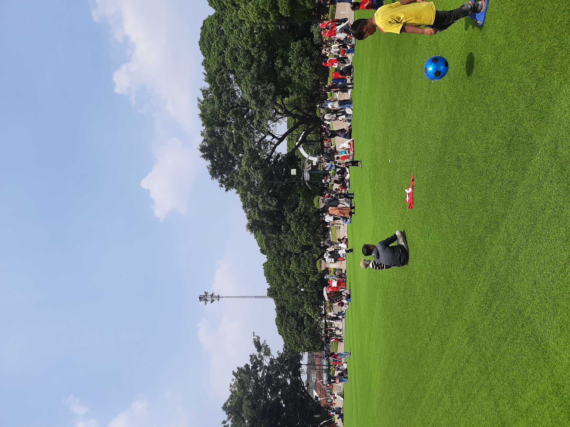 This image depicts a daytime scene in Bandung City, Indonesia, specifically at the Gasibu Field, a popular public space. The field is covered in bright green artificial turf, with a clear blue sky and scattered white clouds overhead. A tall, metallic telecommunications tower is visible in the background. Numerous people are scattered across the field, engaged in various activities.
In the foreground, a young boy in a yellow t-shirt and blue flip-flops is playing with a blue and black soccer ball, which is in mid-air. To his left, a man in a dark grey hooded jacket and light-colored pants is sitting on the grass, facing away from the viewer. Another child, wearing a striped shirt and a light-colored hat, is sitting next to him.
Further back, a diverse group of individuals are spread out, some sitting and conversing, others standing and walking. There are picnic blankets and bags visible amongst the seated people. Lush green trees form a dense canopy in the middle ground and background, with some buildings and structures peeking through the foliage. The overall atmosphere is one of leisure and community gathering on a sunny day.
