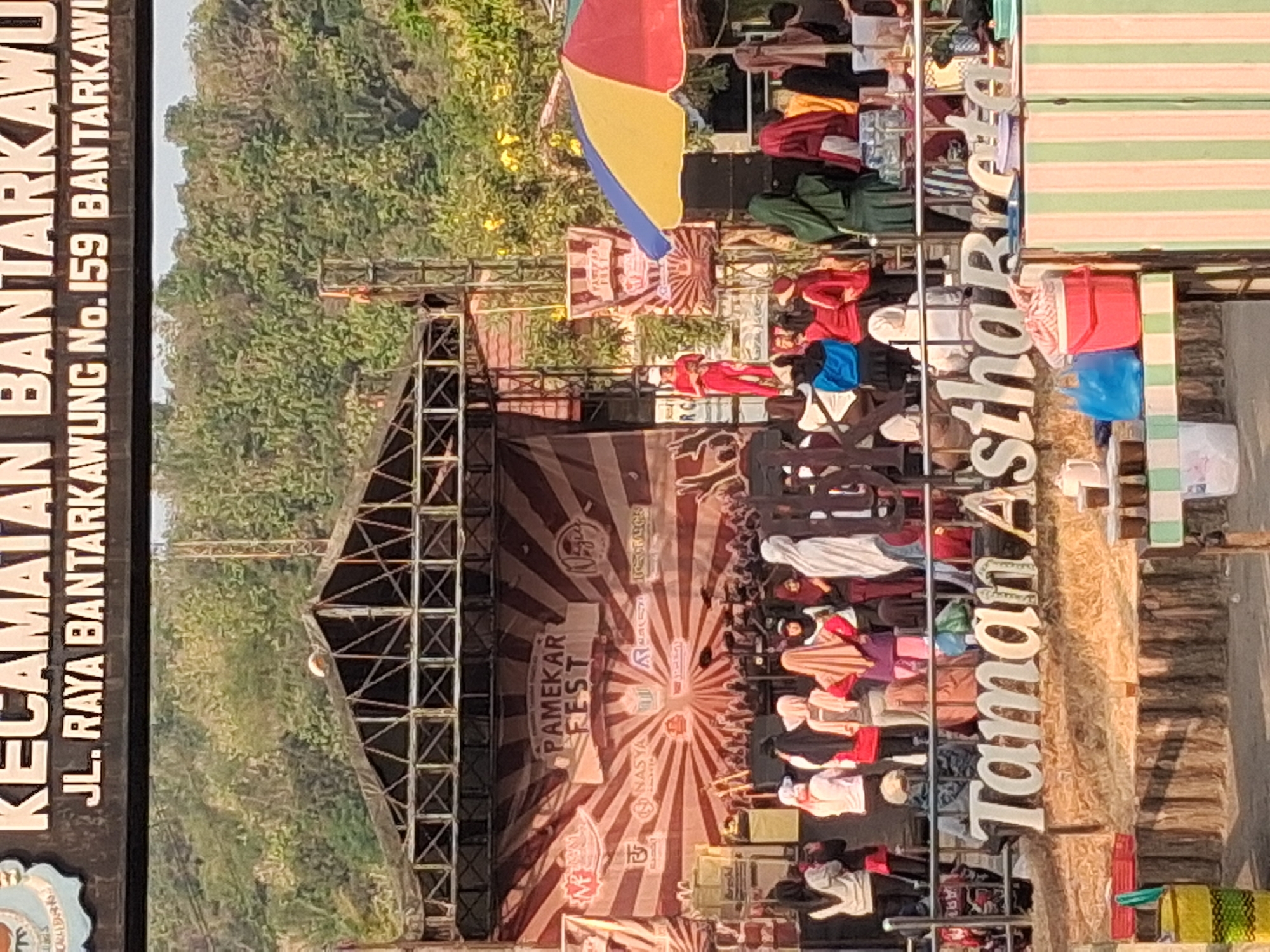 In Bantarkawung, Indonesia, an outdoor event is underway with a stage set up for a performance. A banner advertising "PAMEKAR FEST" hangs on the stage backdrop, featuring a radial striped pattern and various logos. A crowd of people, many wearing headscarves, are gathered in front of the stage, some appearing to be performers or attendees. Among the crowd, a person in a red outfit stands on a raised platform. To the right of the stage, a colorful umbrella shades a section of the area. In the foreground, large white letters spell out "Taman Astha Breta," indicating a park or recreational area. Several tables are set up with goods, including what appear to be drinks and food items, suggesting a marketplace or food stalls. The environment appears to be hilly and green, with trees and vegetation visible in the background.