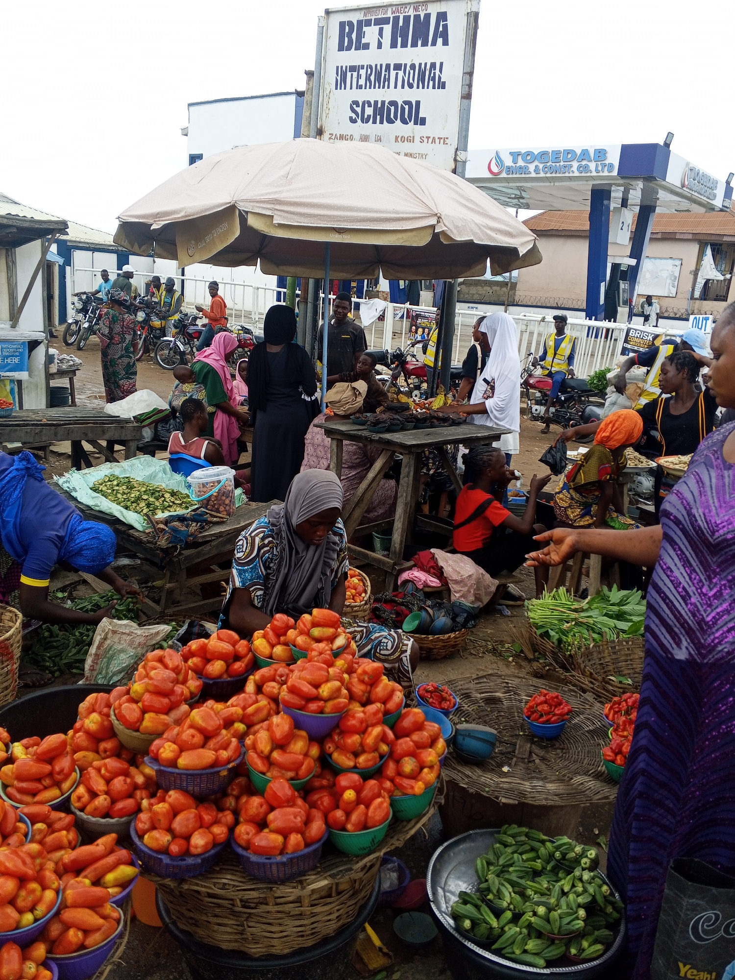 This is a medium shot of an outdoor market in Zango-ndaji, Nigeria, with a gas station visible in the background. The scene depicts a bustling marketplace with numerous people engaged in commerce. Several stalls are set up, displaying a variety of produce including tomatoes and okra. People are seen interacting, with some appearing to be vendors and others customers. Motorcycles are also present, indicating a common mode of transport in the area. The overall environment appears to be a typical Nigerian market scene.
