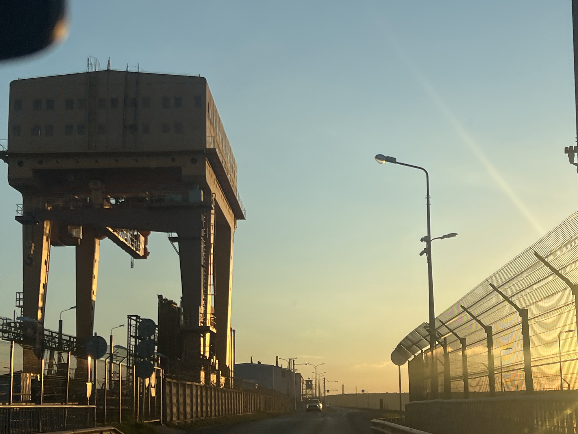 The image depicts a massive, yellow gantry crane situated in an industrial area, likely on the outskirts of Chaikovsky, Russia, as suggested by the presence of similar industrial structures and a characteristic clear sky typical of the region. The sun, low on the horizon, casts a warm, golden light across the scene, creating long shadows and highlighting the metallic textures of the industrial equipment. To the right of the crane, a chain-link fence with a curved top stretches into the distance, also illuminated by the setting sun. A road is visible leading away from the viewer, with a car driving in the distance. Several streetlights are positioned along the road and near the fence. The sky is a gradient of pale blue and warm yellow, indicating either dawn or dusk. The overall impression is one of industrial activity set against a tranquil natural backdrop.