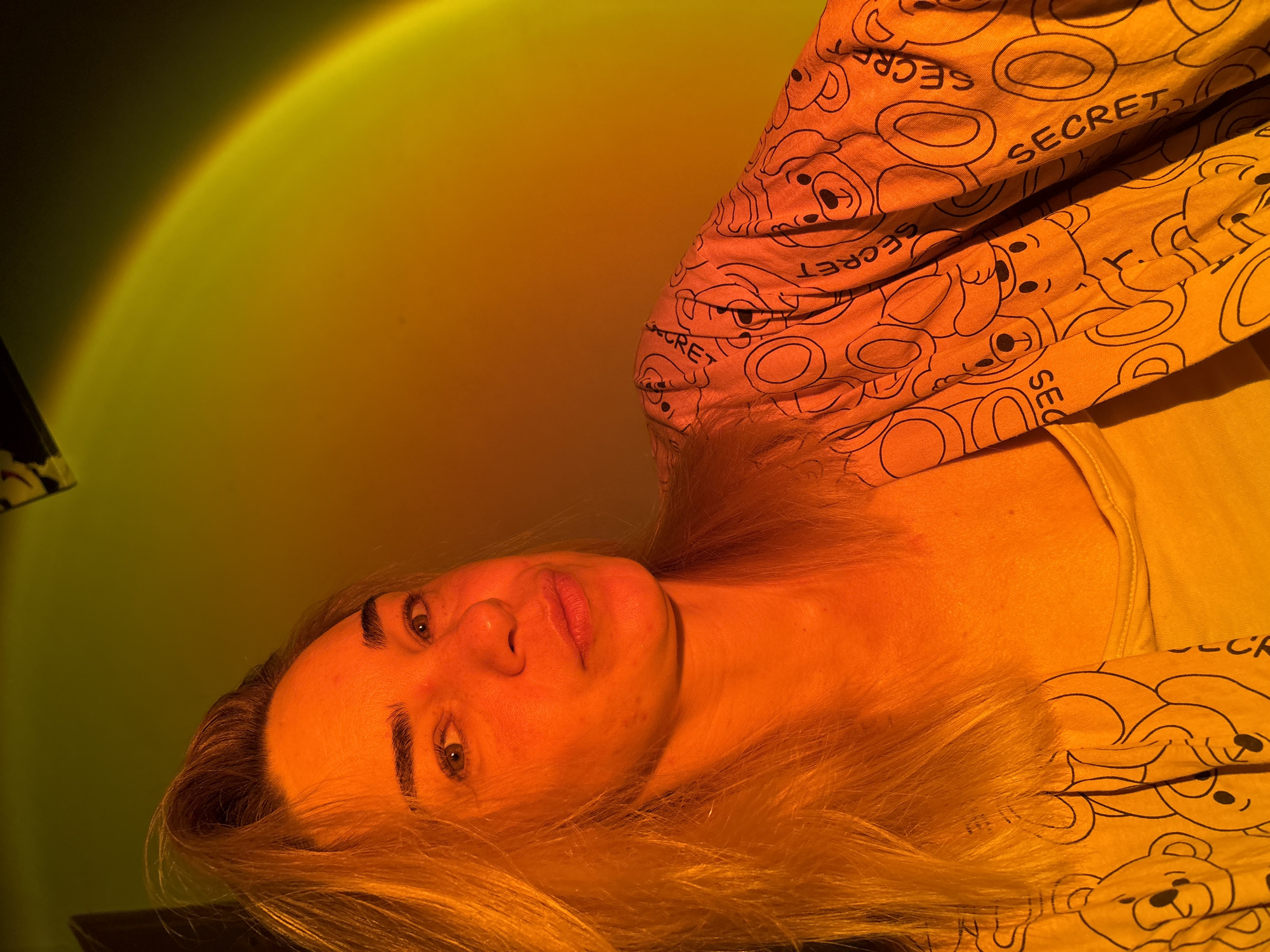 A close-up, waist-up shot captures a young woman with blonde hair bathed in warm, orange and yellow light. She is looking towards the camera with a neutral expression. Her hair is pulled back slightly, with some strands falling forward. She is wearing a light-colored tank top visible beneath an open, long-sleeved garment with a repeating pattern of cartoon teddy bears and the word "SECRET" printed in black. The background is a gradient of yellow and orange, with a hint of a light source creating a circular halo effect. The scene appears to be indoors, likely a home environment, and no specific location in Sinyavino, Russia is identifiable from the visual information provided.