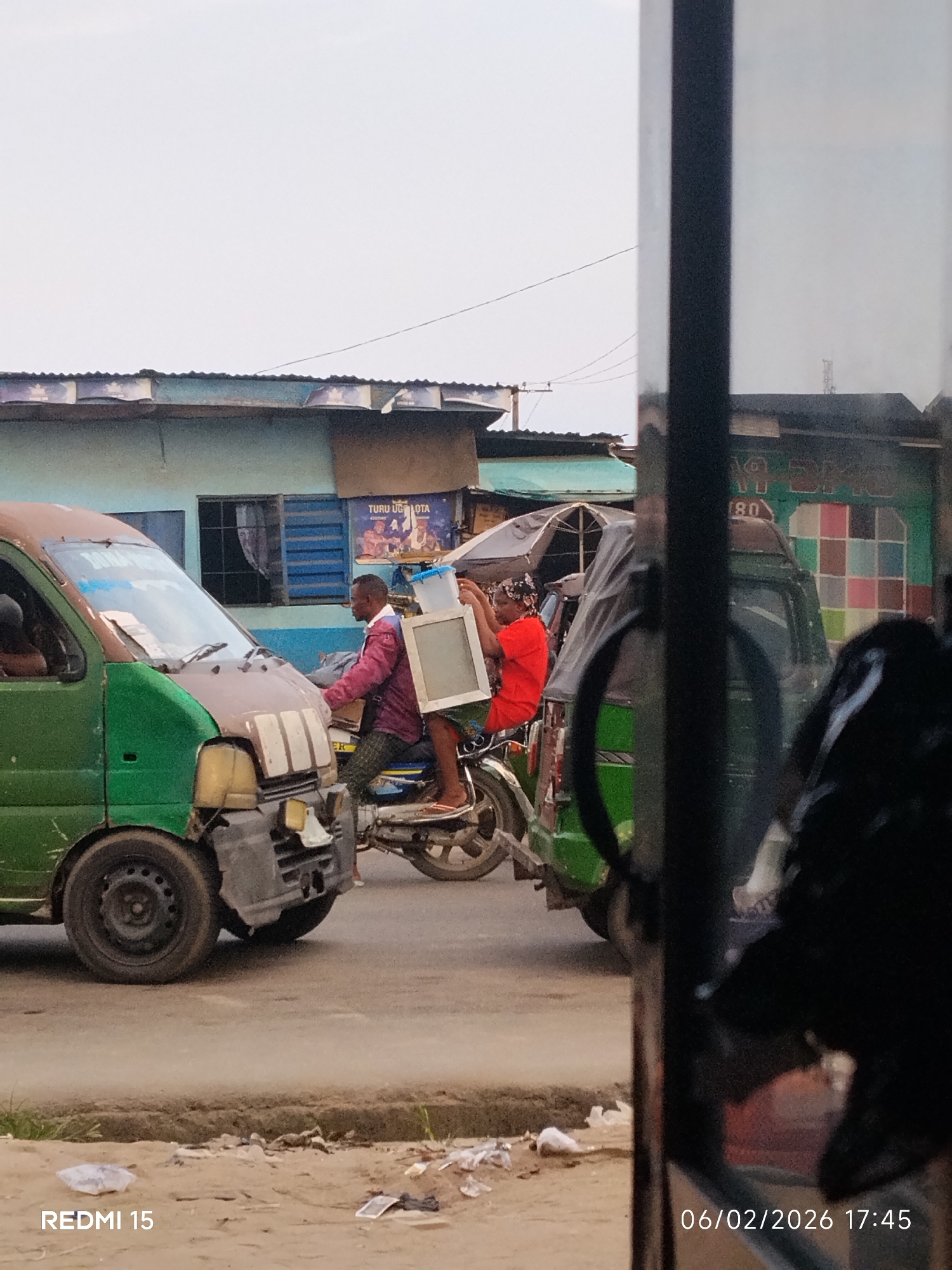This image captures a street scene in Aba, Nigeria, at 17:45 on 06/02/2026, as recorded by a Redmi 15 device. The photograph is taken from behind a prominent dark vertical object in the foreground, partially obscuring the right side of the frame. In the left foreground, a green and brown utility van with a damaged front bumper and exposed components is visible, with a driver's arm seen inside. Central to the image, a man wearing a red long-sleeved shirt and patterned trousers drives a motorcycle. A woman, dressed in a red short-sleeved top and a patterned headwrap, rides as a passenger on the same motorcycle, holding a large, rectangular, light-colored framed object and a smaller, clear plastic container with a blue lid. To the right, another green vehicle, possibly a mini-bus, is partially visible, covered by a dark tarp, with a building behind it featuring a wall painted with a multicolored checkered pattern. In the background, buildings with corrugated roofs are present, one displaying "TURU UGOTA" on a blue sign. Overhead, power lines traverse the pale sky. The unpaved ground in the immediate foreground is dusty and littered with various debris.