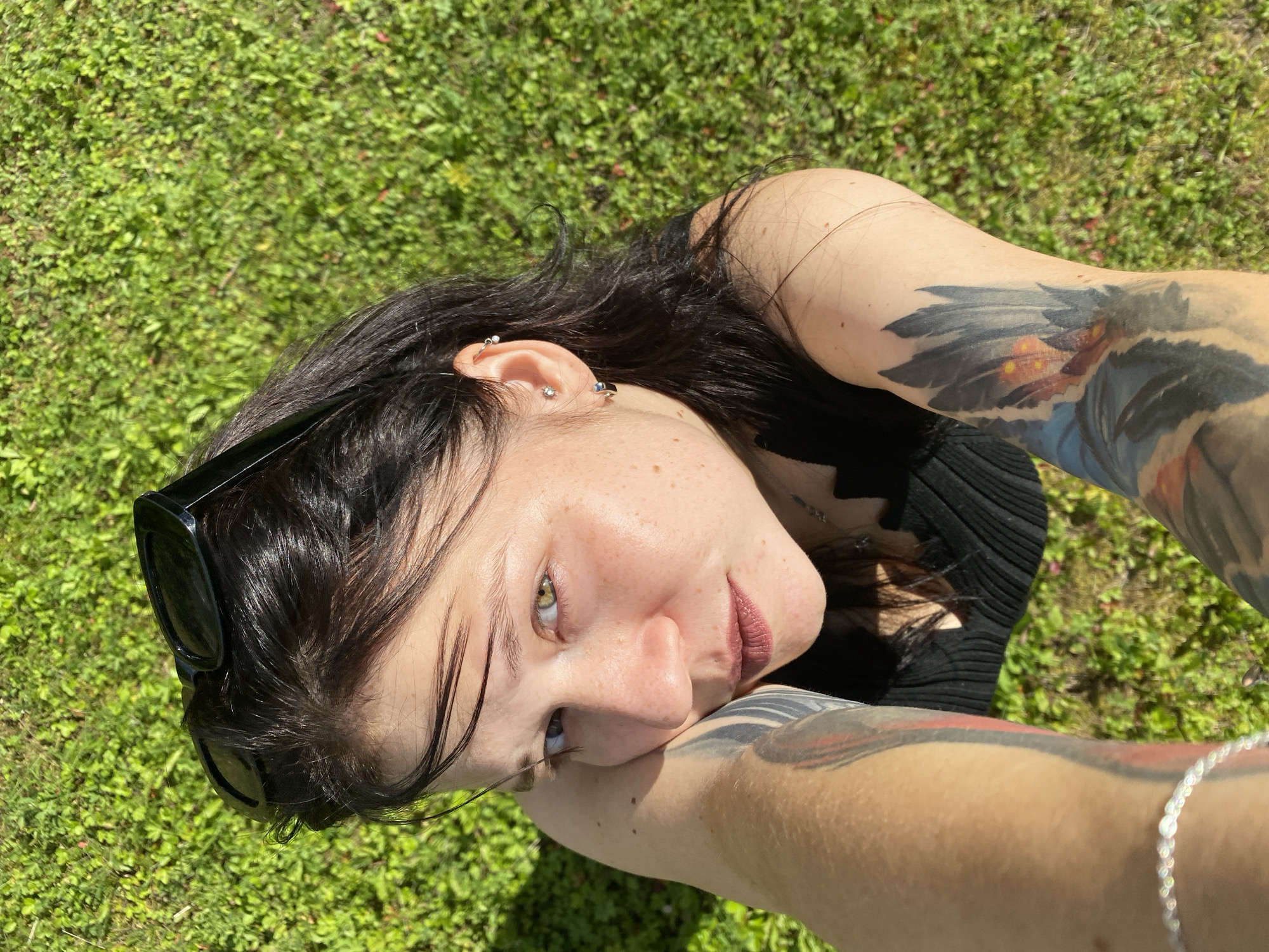 The image is a high-angle, outdoor selfie of a young woman with dark hair and light skin, lying on a patch of green grass. She is looking directly at the camera with a neutral expression. Her left arm is propped up, supporting her head, and is covered in tattoos. She has a dark top on, and sunglasses are perched on top of her head. The background is blurred green grass, indicating an outdoor setting. There are no discernable landmarks or elements that would place the image specifically in the city of Fokino, Russia.