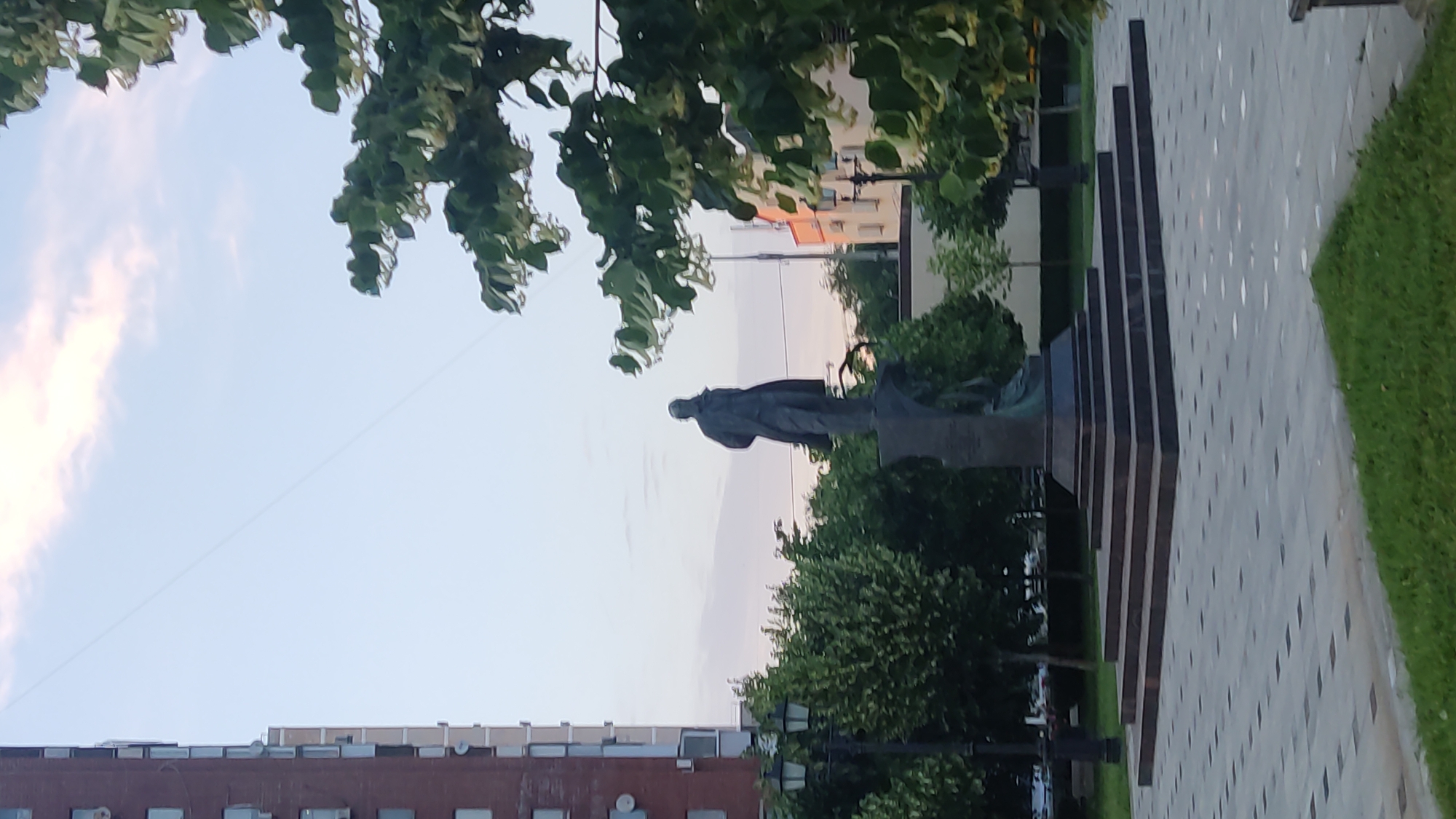 The image shows a monument to Alexei Nikolaevich Kulinich, located in the city of Krasnodar, Russia. The monument features a bronze statue of Kulinich, standing on a tiered pedestal. The statue is dark in color and depicts a man in a coat, looking to the left. The pedestal is made of dark stone and is surrounded by stone steps. The monument is situated in a park-like setting, with trees and green grass visible in the background. A tall, brick building with many windows is to the left of the monument. The sky is visible above, with some clouds. The ground in front of the monument is paved with grey tiles in a geometric pattern.