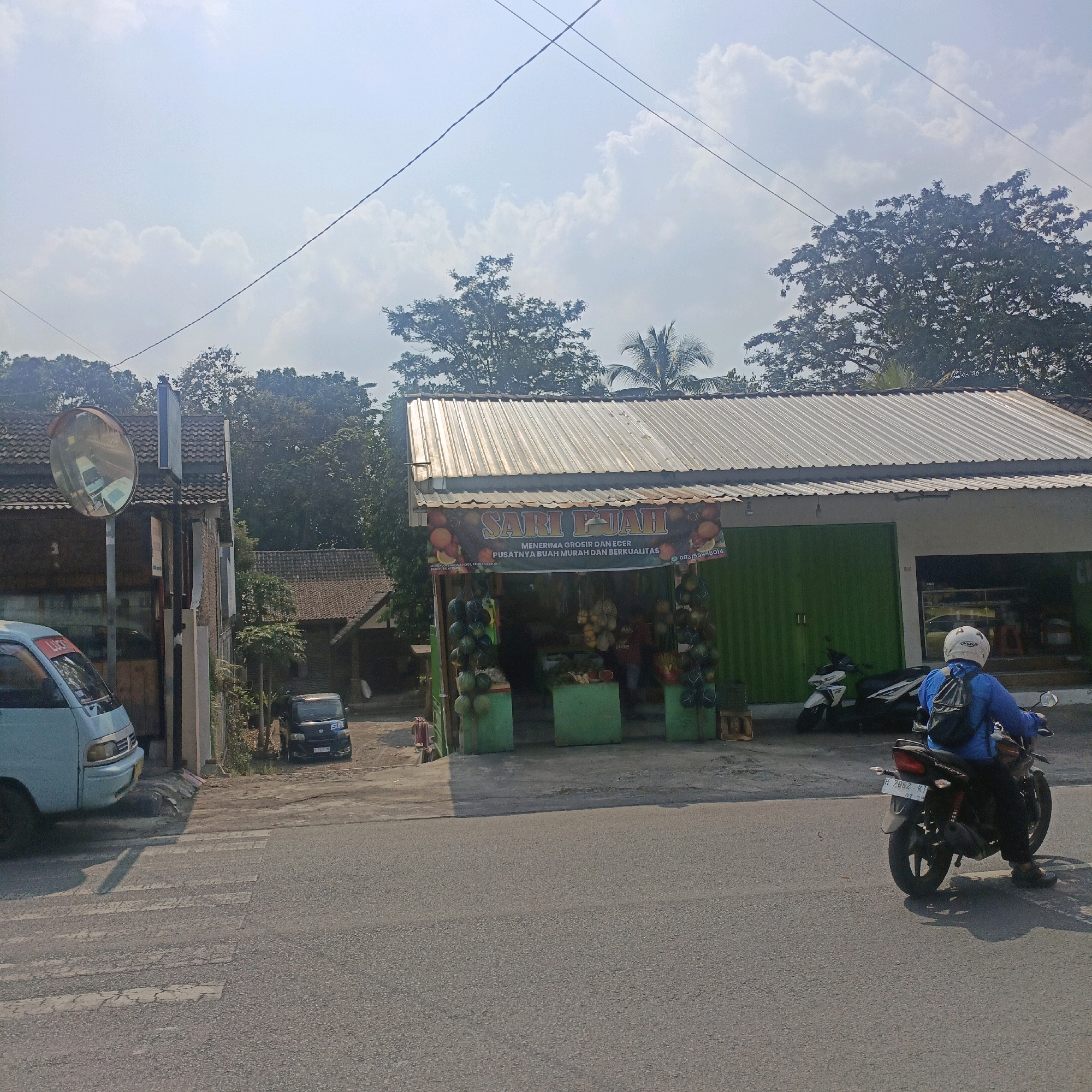 The image shows a street scene in Salatiga, Indonesia, on a bright, sunny day. In the foreground, a paved road with a pedestrian crossing is visible. To the left, a light blue van is parked partially on the side of the road. Across the street, a fruit stand with a corrugated metal roof is set up. The stand displays a variety of fruits, including melons and what appear to be durians, arranged on green display units. A banner above the stand reads "SARI BUAH," indicating it is a fruit shop. To the right of the fruit stand, a person wearing a blue shirt and a white helmet is riding a motorcycle away from the camera. The motorcycle has a license plate with the characters "H 2062 KI". In the background, there are buildings with tiled roofs and lush green trees. A convex mirror is mounted on a pole near the van. Power lines are visible crisscrossing the sky.