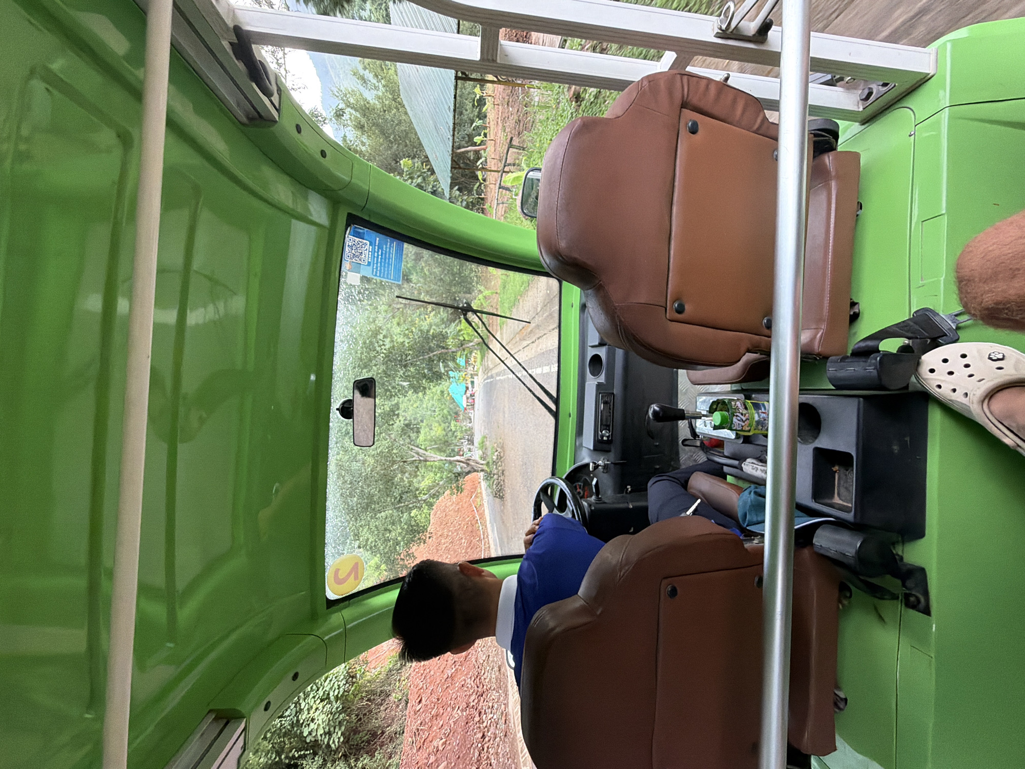 A man is driving a green, open-air golf cart down a paved road in a rural area. The cart has two rows of brown leather seats. The driver, wearing a blue polo shirt and dark pants, is seated in the front left. The road is lined with trees and foliage, and a red, rocky embankment is visible to the left. A green roofed structure with rows of small plants is visible on the right side of the road. The weather appears to be overcast or rainy, as indicated by the droplets on the windshield. The scene is captured from the perspective of a passenger in the back seat. This location is identified as the city of Khanh Vinh, Vietnam.