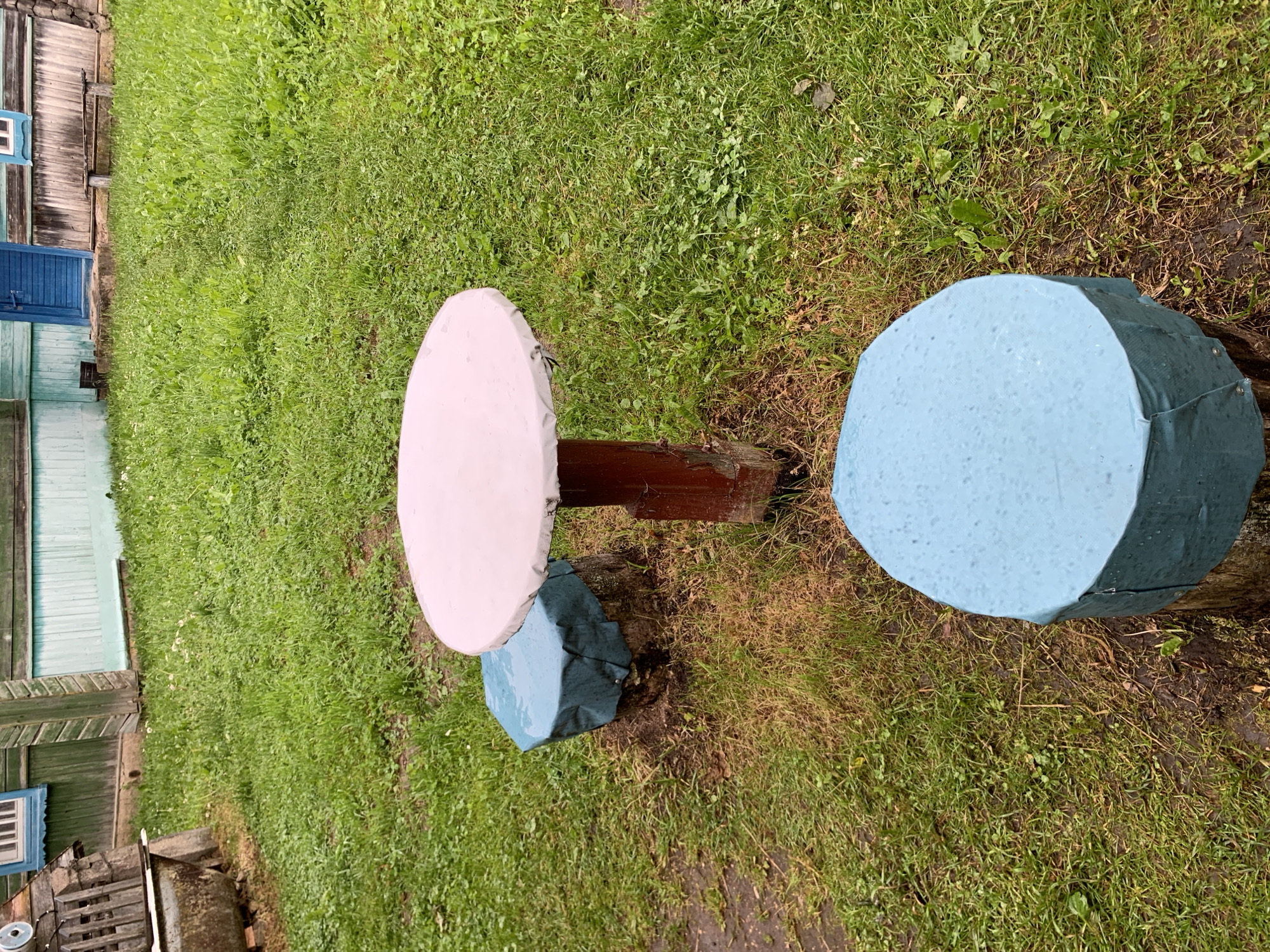 The image shows a circular white-covered table with a red central post standing on a grassy yard. Two round blue-covered stumps are positioned as seating arrangements, one to the left and one in the foreground. The background features a wooden building with green and teal siding and a blue door, possibly a house or shed. The yard is covered in green grass with some brown patches and scattered small white flowers. The scene appears to be outdoors, in a rural setting, possibly the village of Vydza in Belarus.