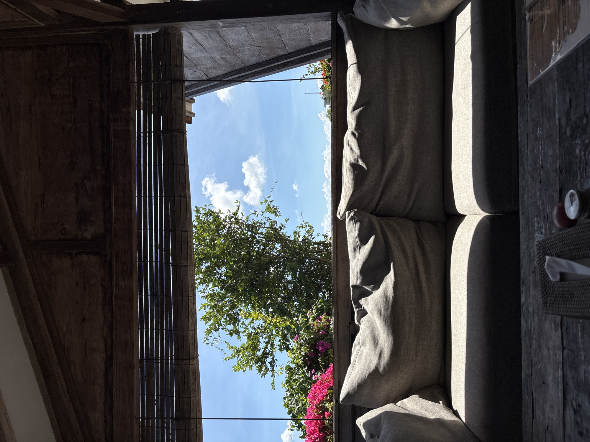 An outdoor seating area in Kuta Selatan, Indonesia, features a large gray couch with two prominent cushions, bathed in sunlight creating stark shadows. In the foreground, a rustic wooden table is partially visible, with a woven napkin holder containing a white napkin and two small round objects. Beyond the couch, a slatted bamboo shade partially obscures a view of lush greenery, including bright pink flowers and a large tree against a bright blue sky with scattered white clouds. The setting appears to be part of a covered patio or gazebo, with exposed wooden beams and a weathered wooden wall visible on the right.