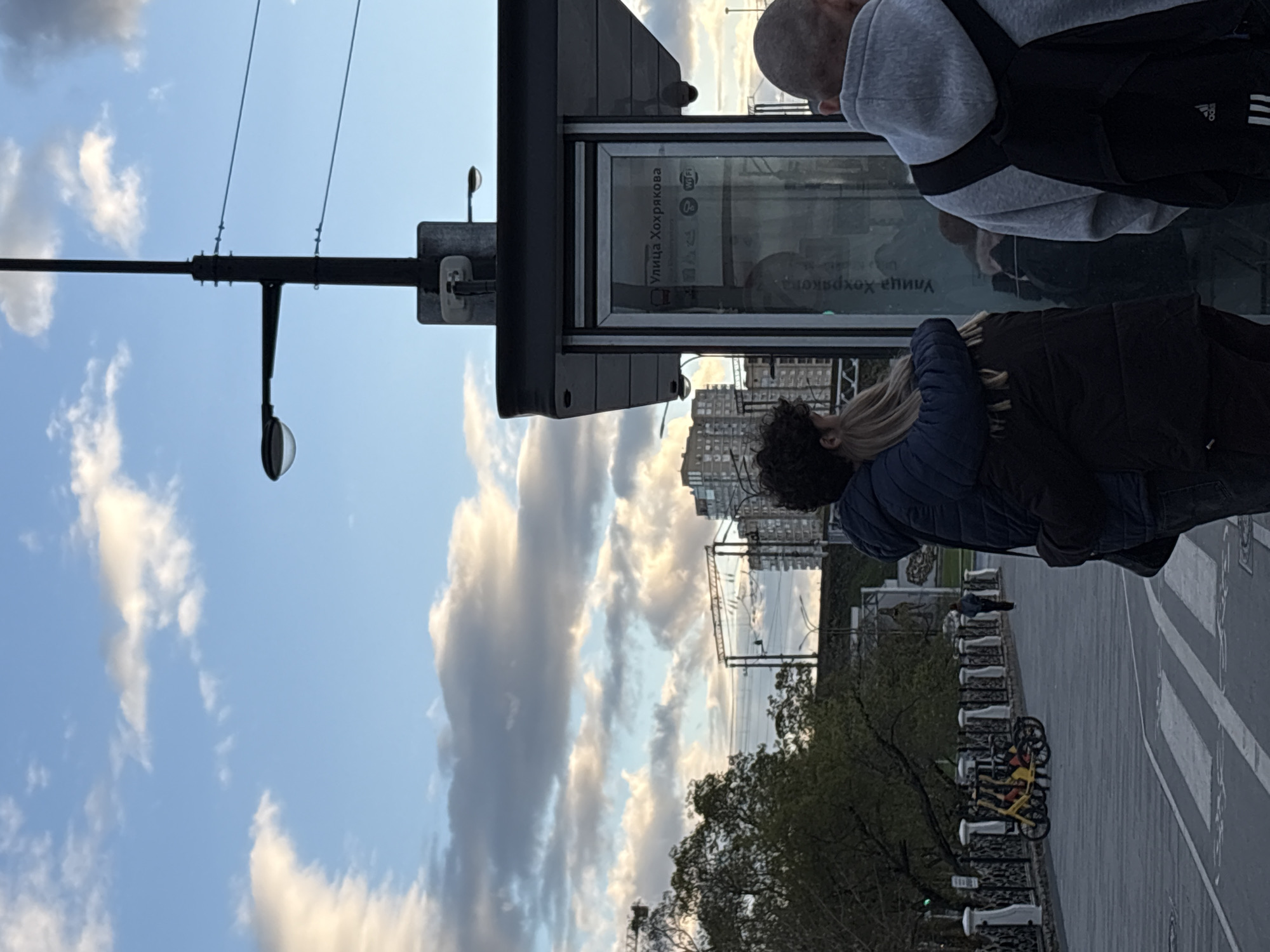 The image shows a bus stop in Perm, Russia. Two people are looking at the bus schedule, which is written in Russian. The bus stop is located on Ulitsa Khohryakova, a street in Perm. There is a bike rack next to the bus stop. Across the street is some greenery with trees, beyond which can be seen a complex of buildings. The sky is blue with scattered clouds. A street light is visible at the left of the frame.