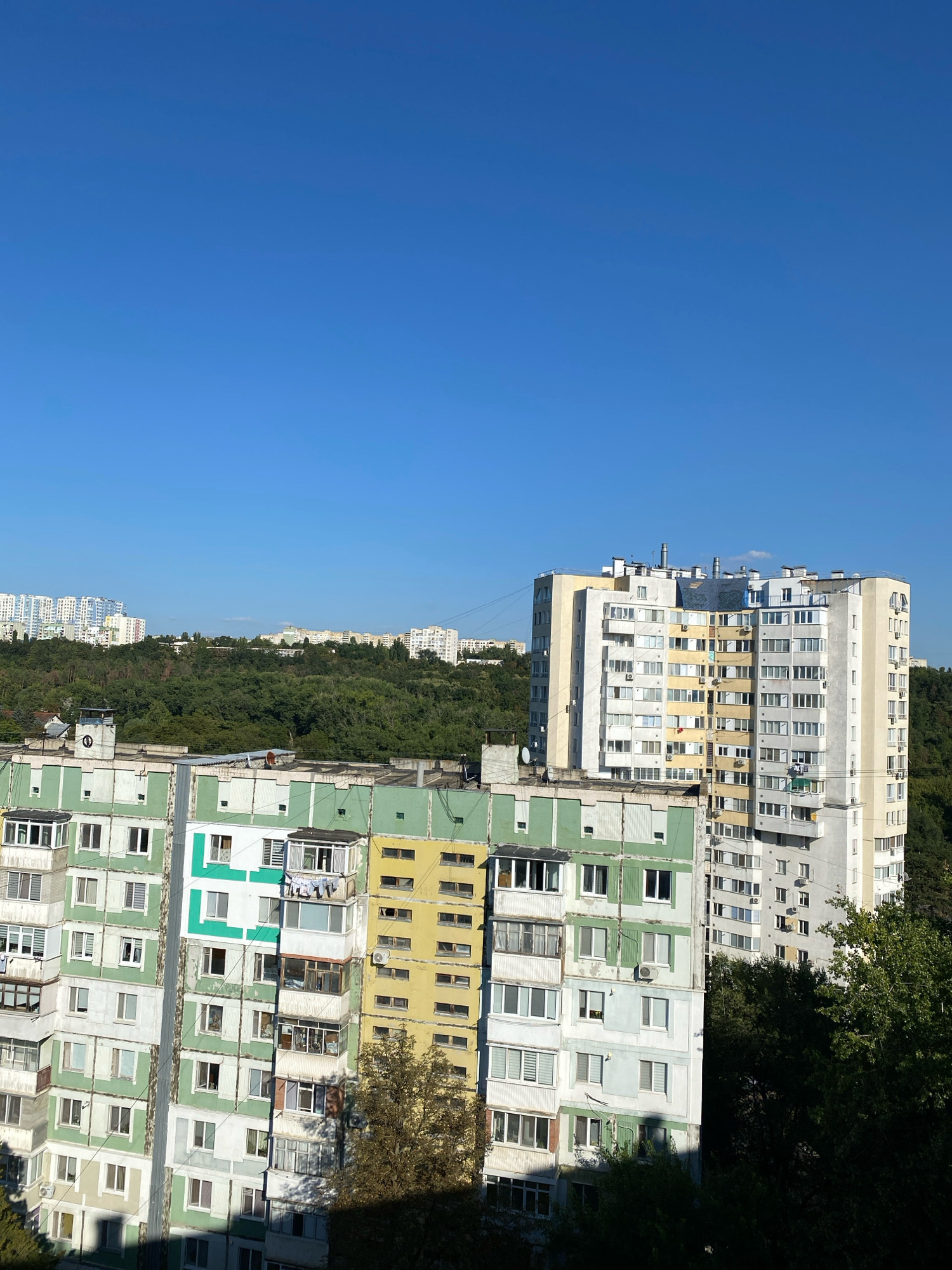 This is an eye-level, medium shot of a residential area in Chișinău, Moldova, on a clear, sunny day. The sky is a vibrant, cloudless blue. In the foreground, a row of apartment buildings with varying facades is visible. The closest building on the left is predominantly white and green, with balconies and numerous windows. To its right, another apartment building displays a mix of pale yellow, green, and white sections. Further in the background, a taller, more modern apartment building stands out with its pale yellow and white exterior, featuring many windows and air conditioning units. Behind the buildings, a dense, green hillside covered in trees stretches across the middle ground, creating a natural backdrop. In the far distance, several other apartment buildings are visible on the horizon, suggesting a sprawling urban landscape. The scene appears to be a typical view of residential blocks in a city, with a blend of older and newer architecture set against a natural landscape. There are no people or animals visible in the image.
