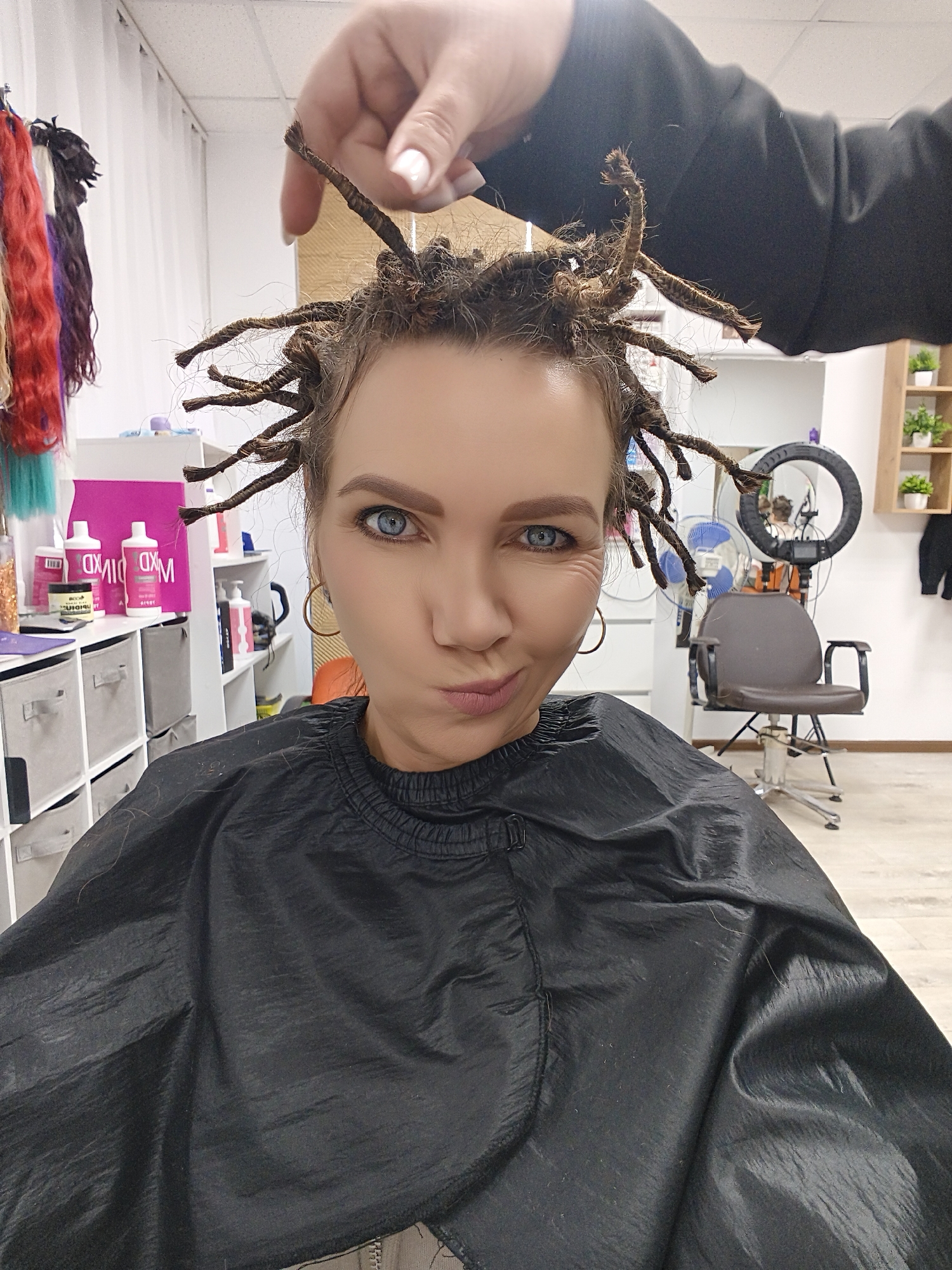 A medium close-up shot captures a woman sitting in a hair salon or studio, looking directly at the camera with a playful, pouting expression. Her striking blue eyes are a focal point, and she has light brown hair styled into numerous dreadlock-like twists that stick out from her head. One hand, presumably belonging to a stylist, is visible at the top of the frame, holding one of the hair twists, indicating an ongoing hair styling process.
The woman is wearing a black, shiny salon cape that covers her shoulders and chest. A gold hoop earring is visible on her left ear. In the background to the left, a rack displays colorful hair extensions in shades of red, purple, blonde, and teal. Below this, various hair products, including white and pink bottles (some with the text "MDXID" or "MIXED" visible, potentially reversed), are arranged on shelves and a counter, alongside a container of glitter.
To the right of the woman, the setting continues to reveal a professional salon environment. A black, swiveling salon chair with a chrome base is prominent, next to a large, circular LED ring light on a tripod. A white floor fan is also visible, and in the far background, a wooden shelf unit holds small potted green plants. The walls are white, and the floor appears to be a light-colored wood or laminate. The overall impression is that of a relaxed, in-progress hair transformation in a well-equipped studio.