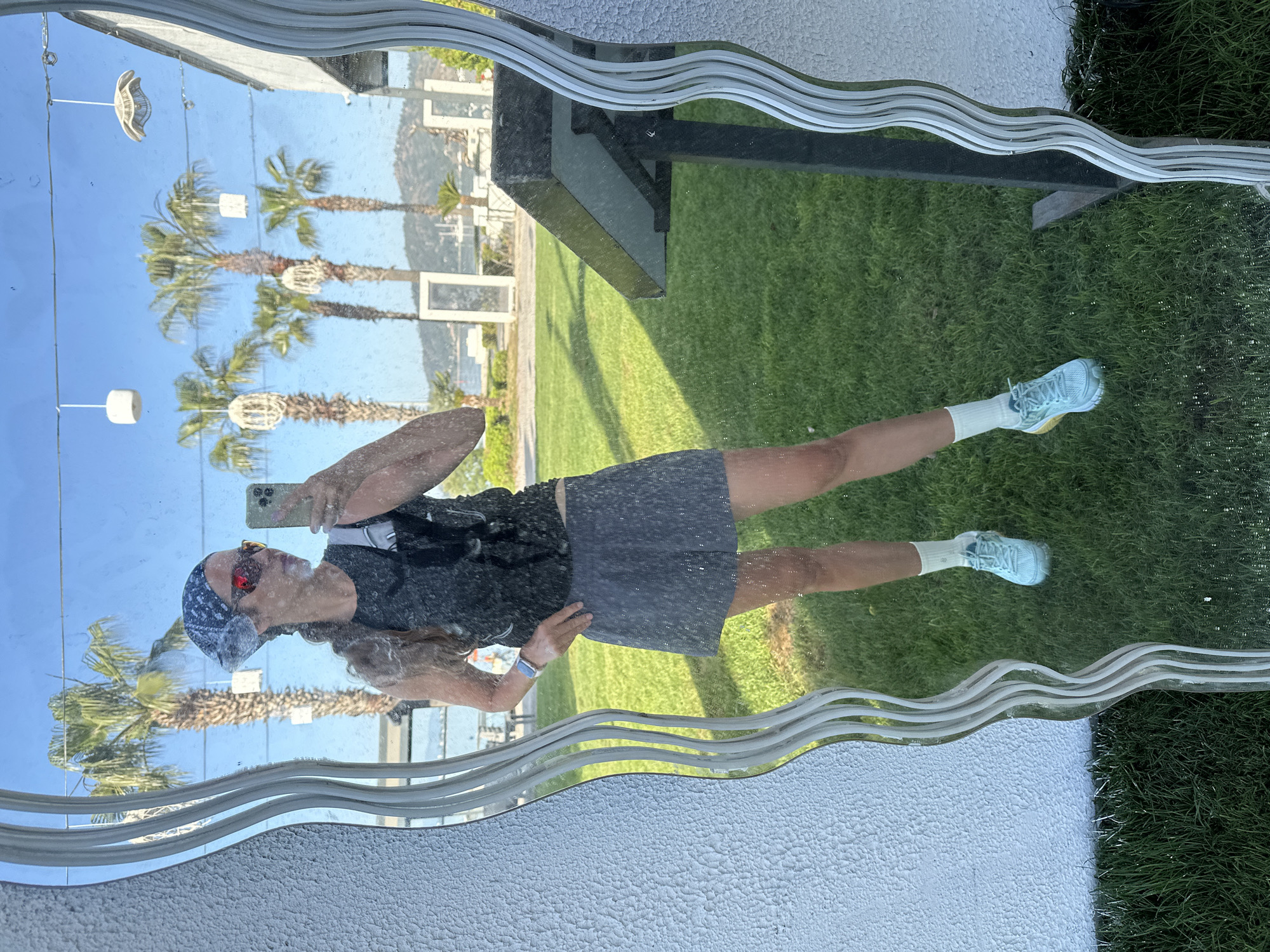 The image shows a person's reflection in a decorative mirror. The person is wearing a dark tank top, gray shorts, white socks, and light blue sneakers. They are also wearing a patterned bandana on their head and sunglasses. They are holding a phone up to take a picture of themself in the mirror. Behind the person, visible in the reflection, are palm trees, white decorations hanging from wires, and a blue sky. Behind the mirror, but also visible in the reflection, is a green lawn and a black structure that could be part of a bench or building. The ground around the mirror appears to be a textured white surface. Given the presence of palm trees and the blue sky, the location is in Fethiye, Turkey.