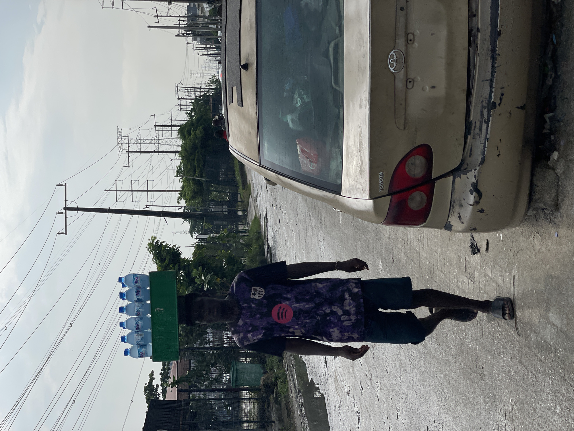 In Alaguntan, Nigeria, a young Black male walks along a wet, paved road, carrying a green crate of bottled water balanced on his head. He is wearing a dark t-shirt with a purple, black, and pink abstract design and black shorts. His feet are clad in black slides. To the right of the frame is the rear of a weathered, tan Toyota sedan, its bumper visibly damaged and scratched. The road appears to have standing water in puddles, suggesting recent rain or poor drainage. In the background, numerous electrical poles and power lines crisscross the sky, indicative of urban infrastructure. Trees and other vegetation are visible on the left side of the road, with a fence and some structures behind them. The sky is overcast.