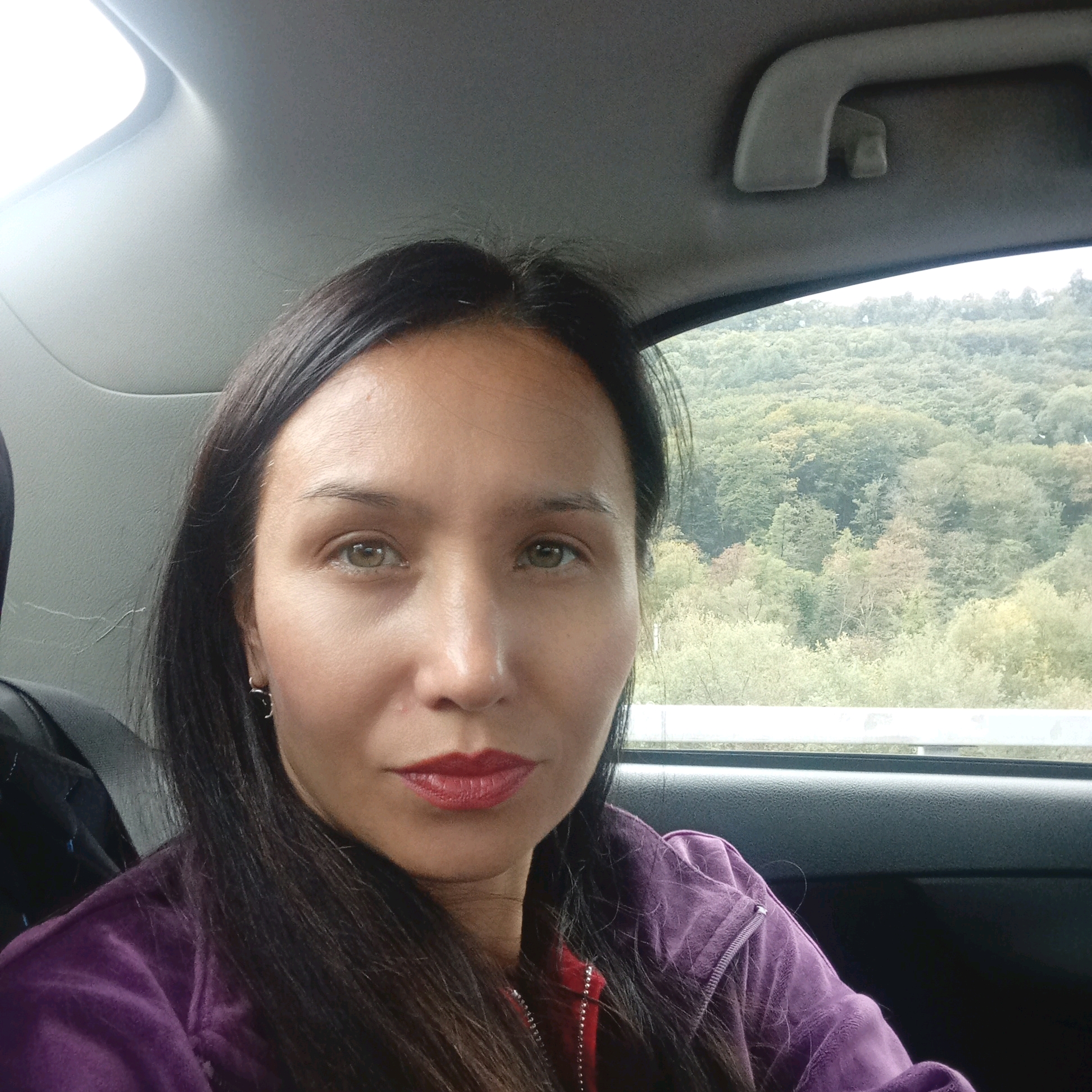 In a selfie taken from inside a vehicle, a woman with dark hair and hazel eyes looks towards the camera. She is wearing a purple velour jacket over a red top. Her lips are painted with red lipstick. The background shows a blurred, green, tree-covered hillside, suggesting a natural environment. The interior of the car, with its headliner and door panel, is visible. The overall lighting suggests it is daytime.