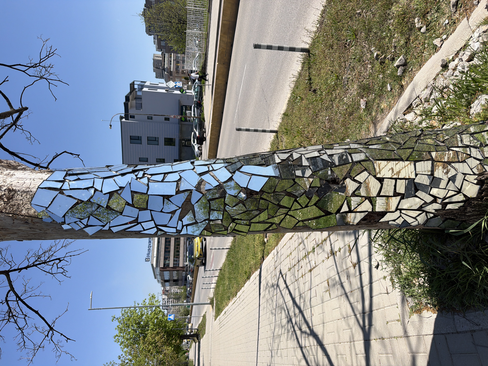 The image captures a distinctive urban art installation in Sofia, Bulgaria, featuring a vertical tree trunk adorned with a mosaic of numerous irregularly shaped mirror fragments. These mirrors cover a significant portion of the tree's bark, creating a fragmented reflection of its surroundings. Above the mosaic, the tree's branches are largely bare against a clear blue sky, though some green foliage is reflected in the mirrors and visible further down the street.
The scene depicts a sunny city street on a bright day. To the left of the mirrored tree, a patterned paved sidewalk stretches, casting distinct shadows. Beyond the sidewalk, various urban buildings are visible, one prominently displaying the "Husqvarna" logo. A blue pedestrian crossing sign can also be seen in the distance. To the right of the tree, a strip of green grass borders an asphalt road with a clear white center line. Two black and white striped bollards stand at the road's edge. Cars are parked and moving along the street in the background, contributing to the urban setting.
The mirror mosaic on the tree creates a dynamic visual effect, reflecting and distorting elements like the blue sky, green grass, and building facades. The bright daylight enhances the reflective quality, making the tree a striking and interactive piece of public art within the city landscape.