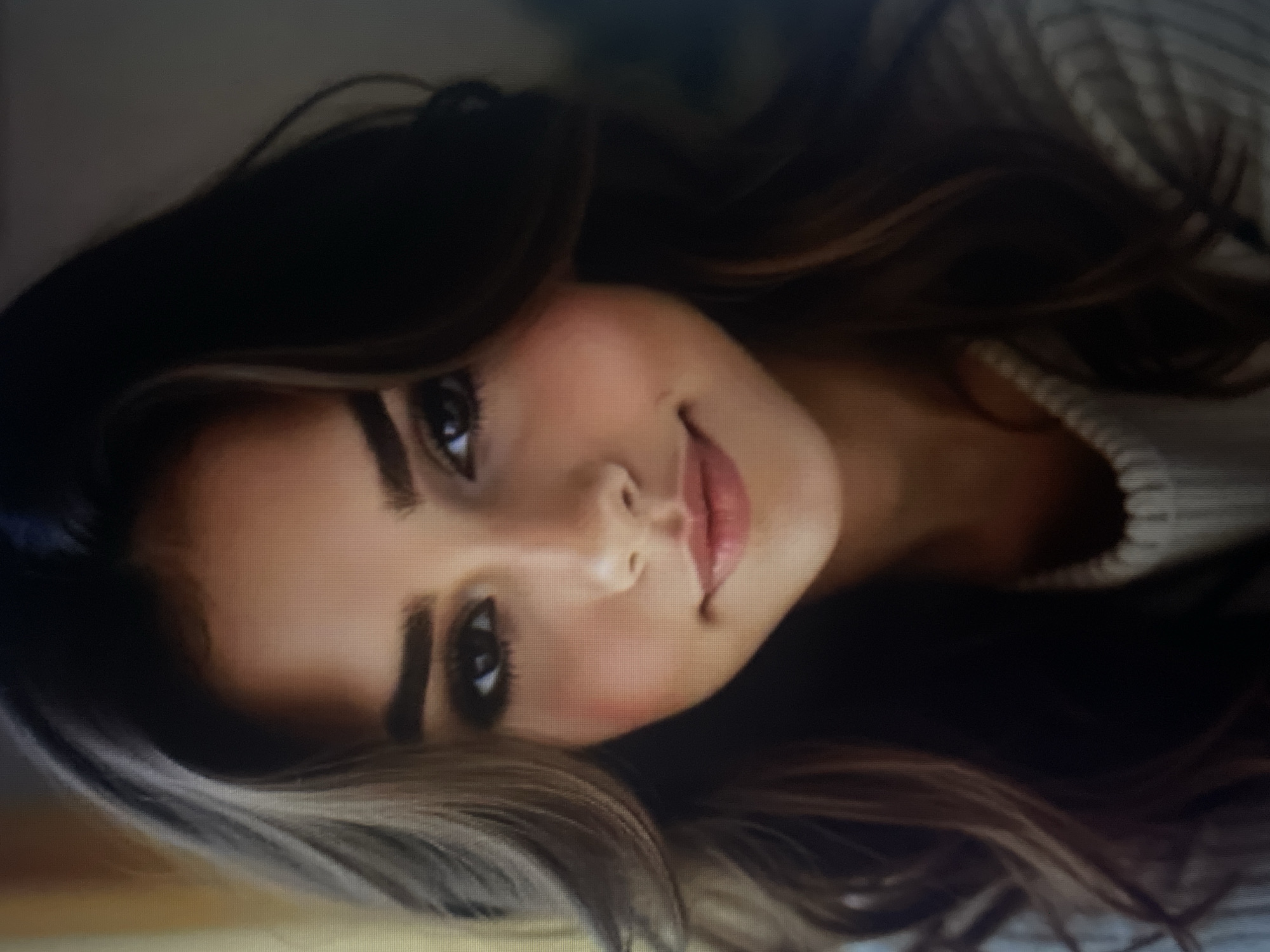 The image is a close-up, studio portrait of a young woman with dark brown hair and fair skin. She is wearing a light-colored, ribbed sweater. The background is blurred and consists of soft, warm tones, suggesting an indoor setting. There is no discernible location in the city of Kołobrzeg, Poland. The woman is looking directly at the camera with a slight smile. Her makeup includes dark eyeliner and mascara, and blush on her cheeks.
